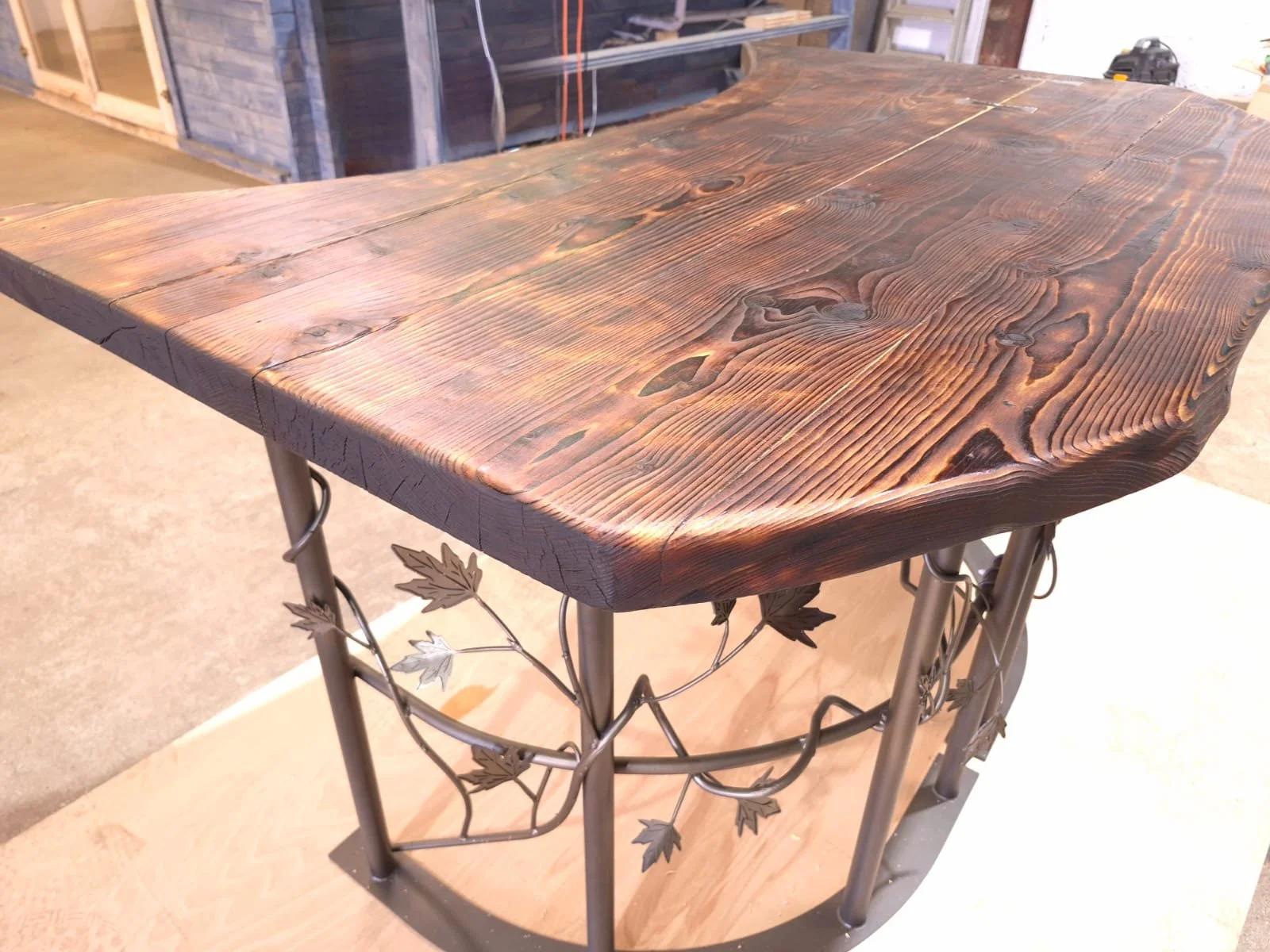 A wooden table with a natural, darkened finish and rounded edges. The table has a metal base with decorative vine-like details.