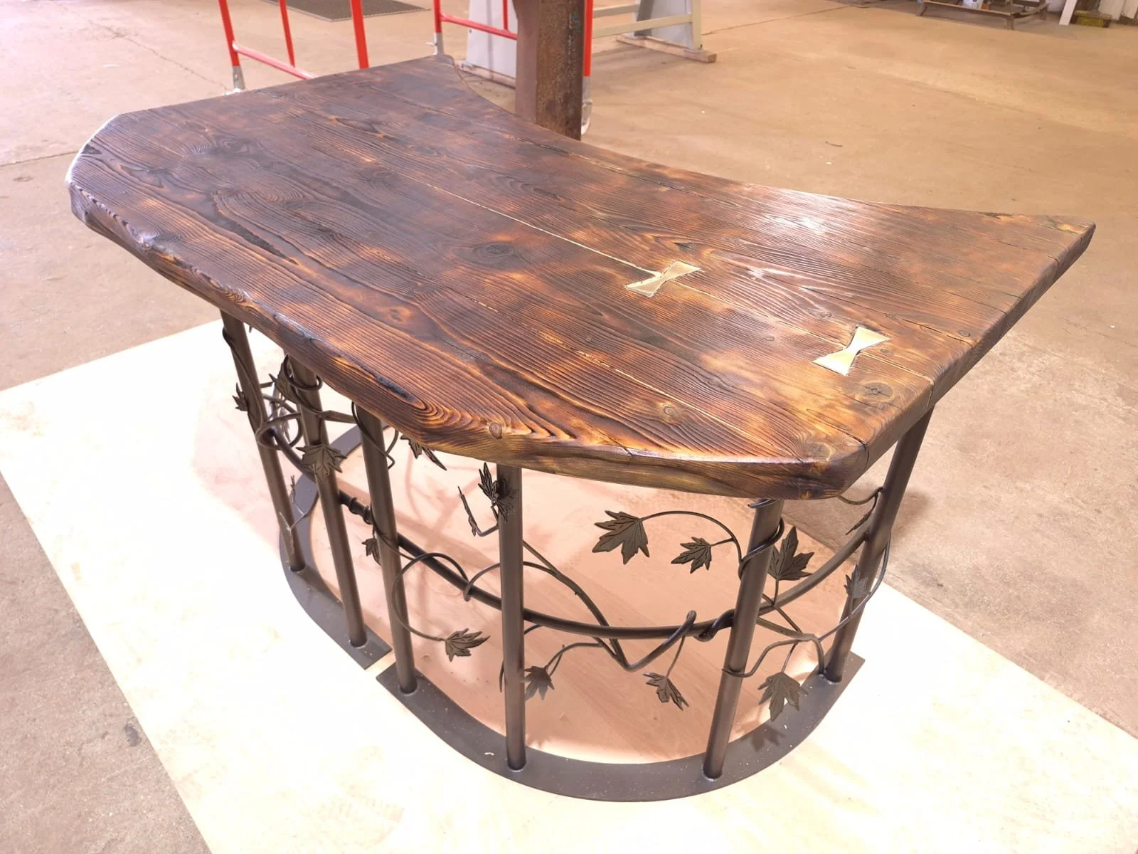 reclaimed top with a scorched finish, supported by a black metal base with leaf and vine decorative details.