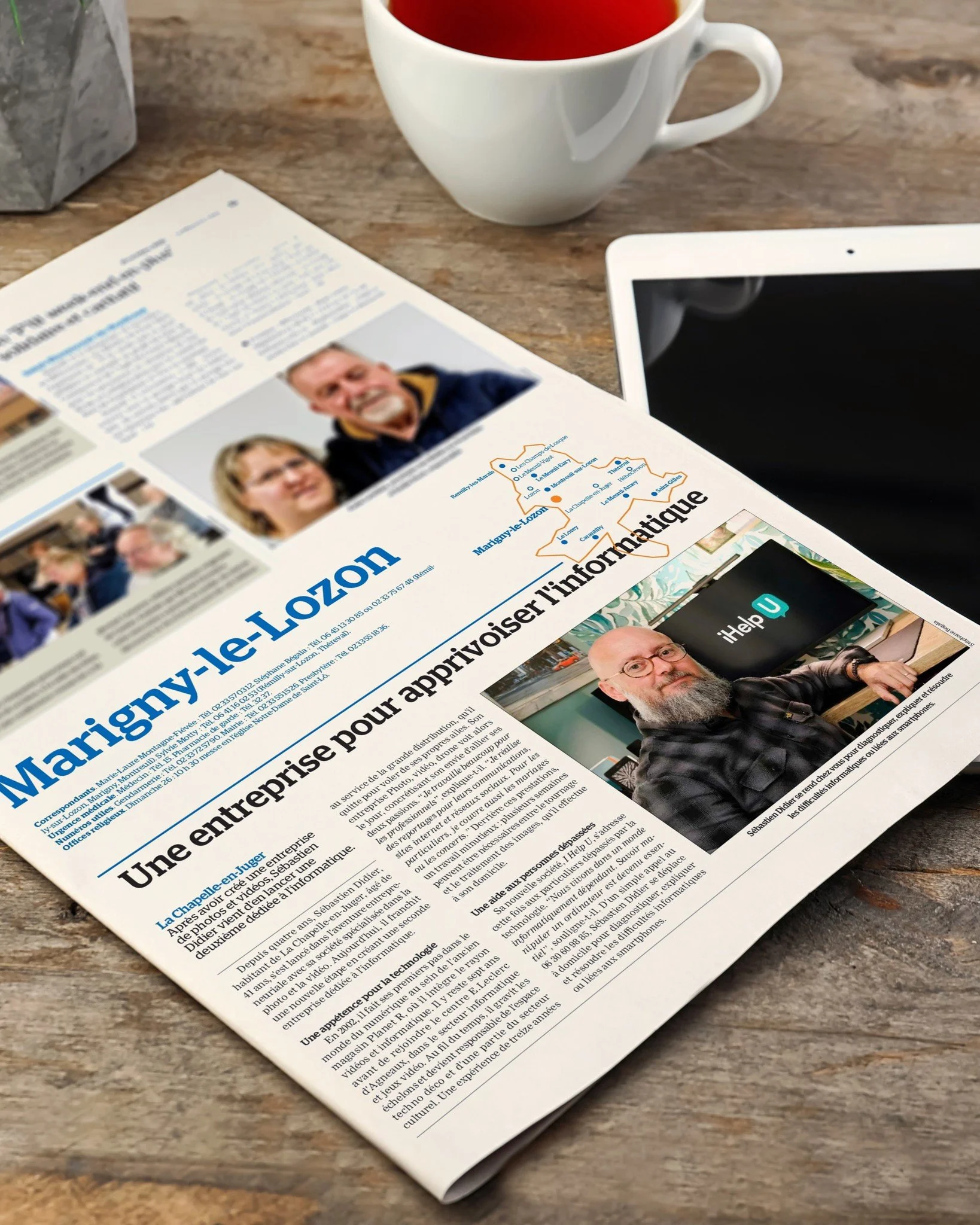 Une page de journal avec un article intitulé "Une entreprise pour apprivoiser l'informatique", une photo d'un homme barbu avec des lunettes devant un écran, une carte de la région de Marigny-le-Lizon, une tasse de thé ou café, un téléphone portable, et un document en arrière-plan sur une surface en bois.