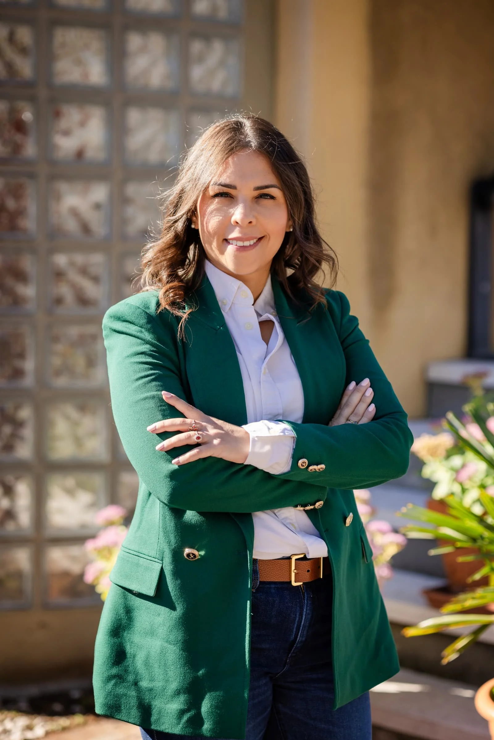 Una donna sorridente con capelli castani corti, vestita con una giacca verde, camicia bianca e jeans, che si trova all'aperto davanti a un muro di mattoni di vetro e piante fiorite.