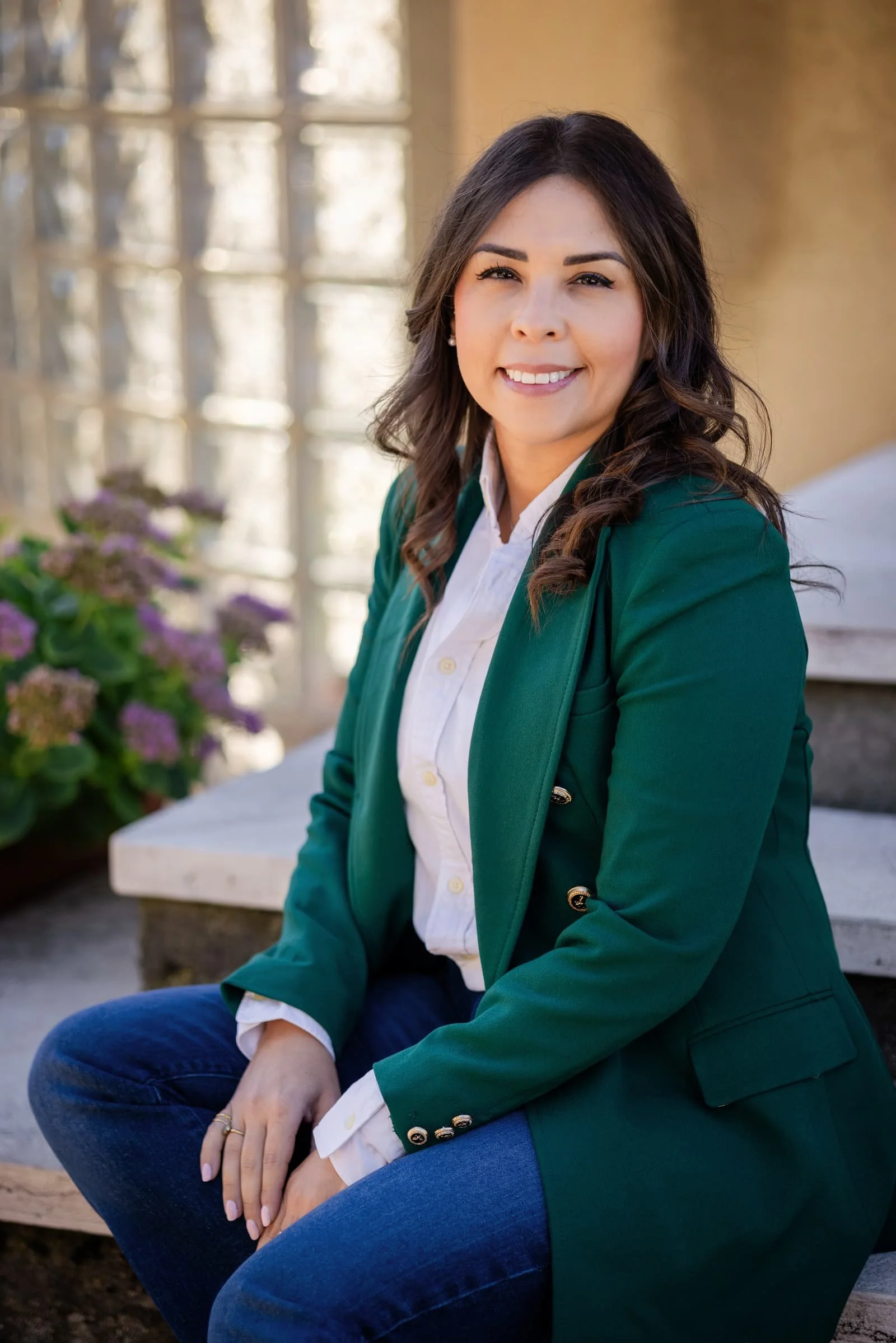 Una donna con capelli riccioli castani chiari, vestita con una camicia bianca e una giacca verde scuro, seduta su una panchina con fiori di orchidea rosa e arbusti sullo sfondo, in un ambiente all'aperto.