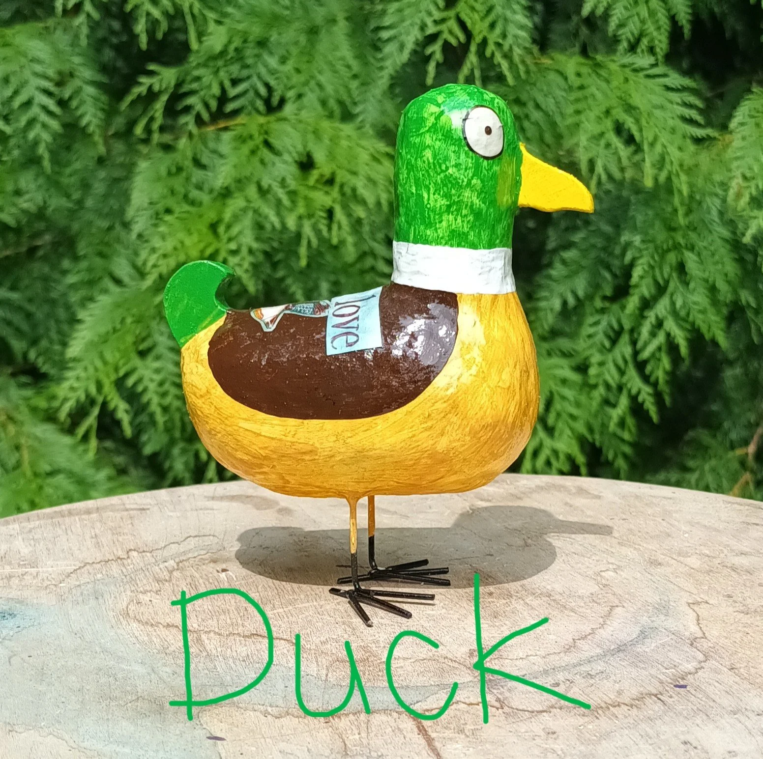 A small Posh Paper Mache mixed media duck waterbird sculpture standing on a wooden plinth against a backdrop of pine trees, photographed in the artists garden.