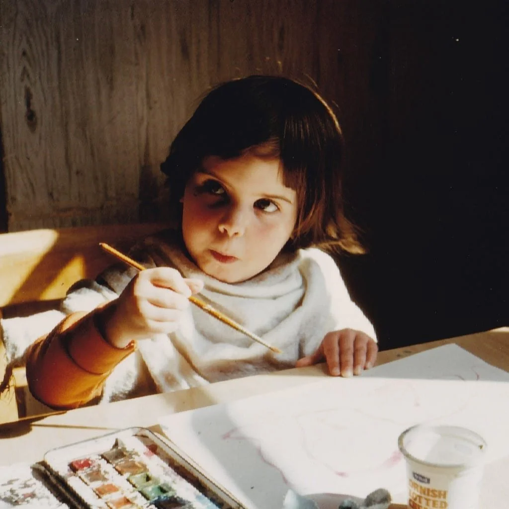A young girl with short dark hair is sitting at a table with a paint set and a glass of milk, holding a paintbrush, and appears to be painting.
