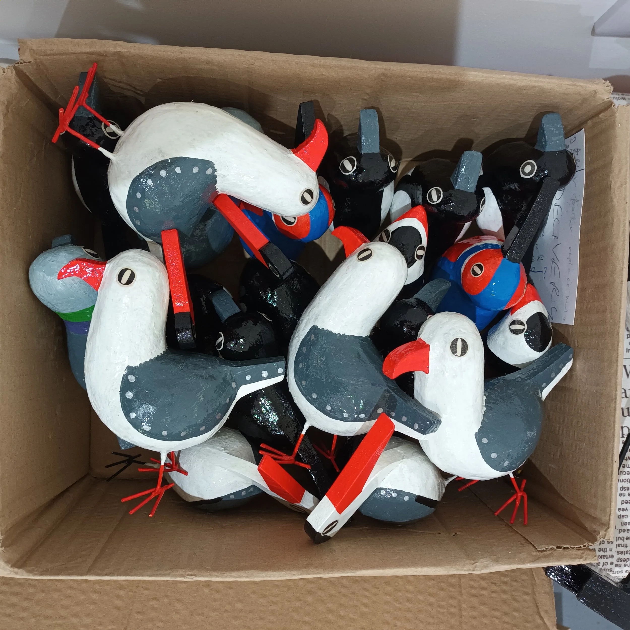 A cardboard box filled with wooden bird posh paper mache sculptures including seagulls and crows painted in various colors, primarily black, white, red, and blue, depicting puffins and other seabirds.