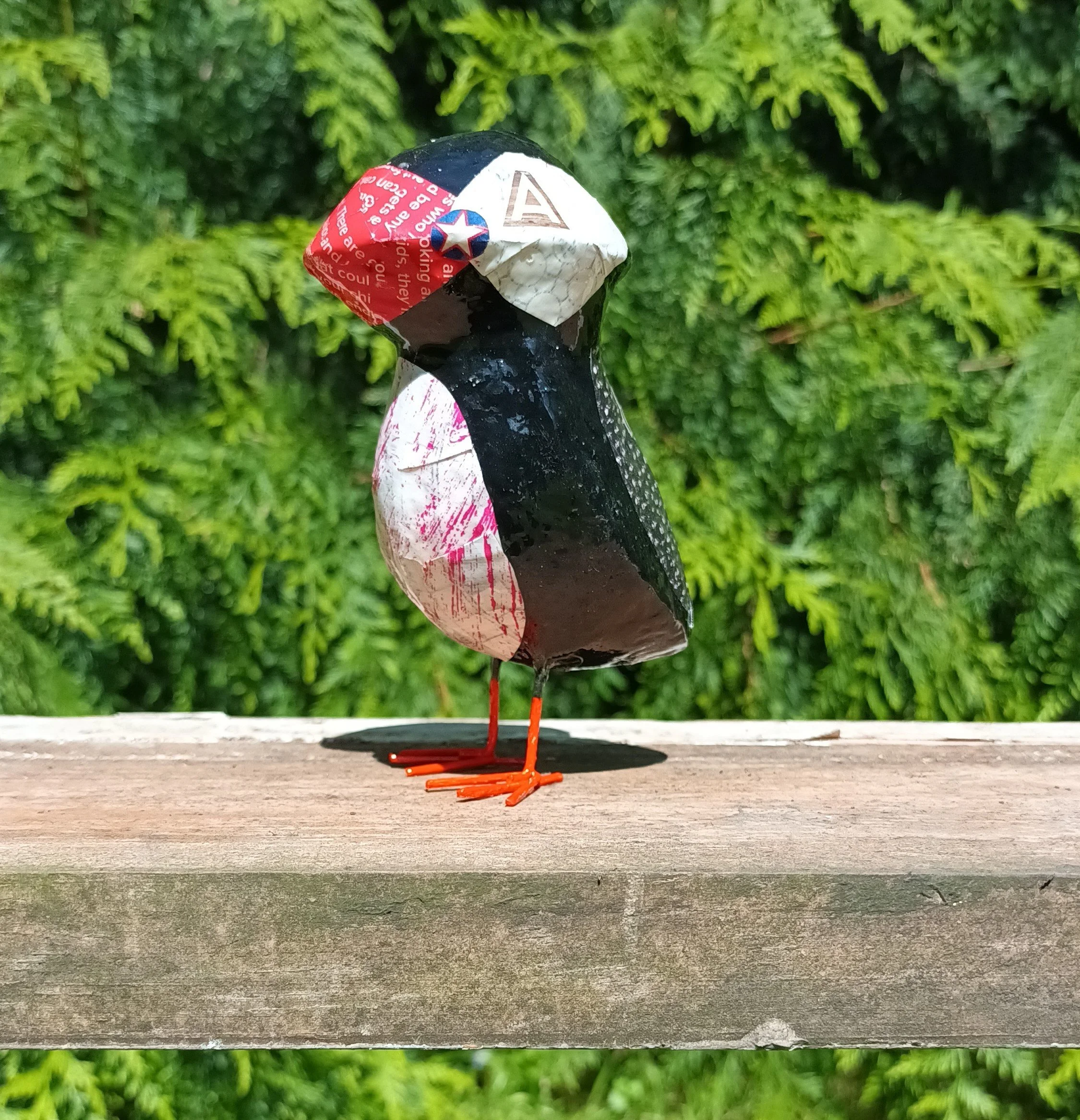 A handmade bird sculpture made from recycled paper and metal, sitting on a wooden surface against a green leafy background.