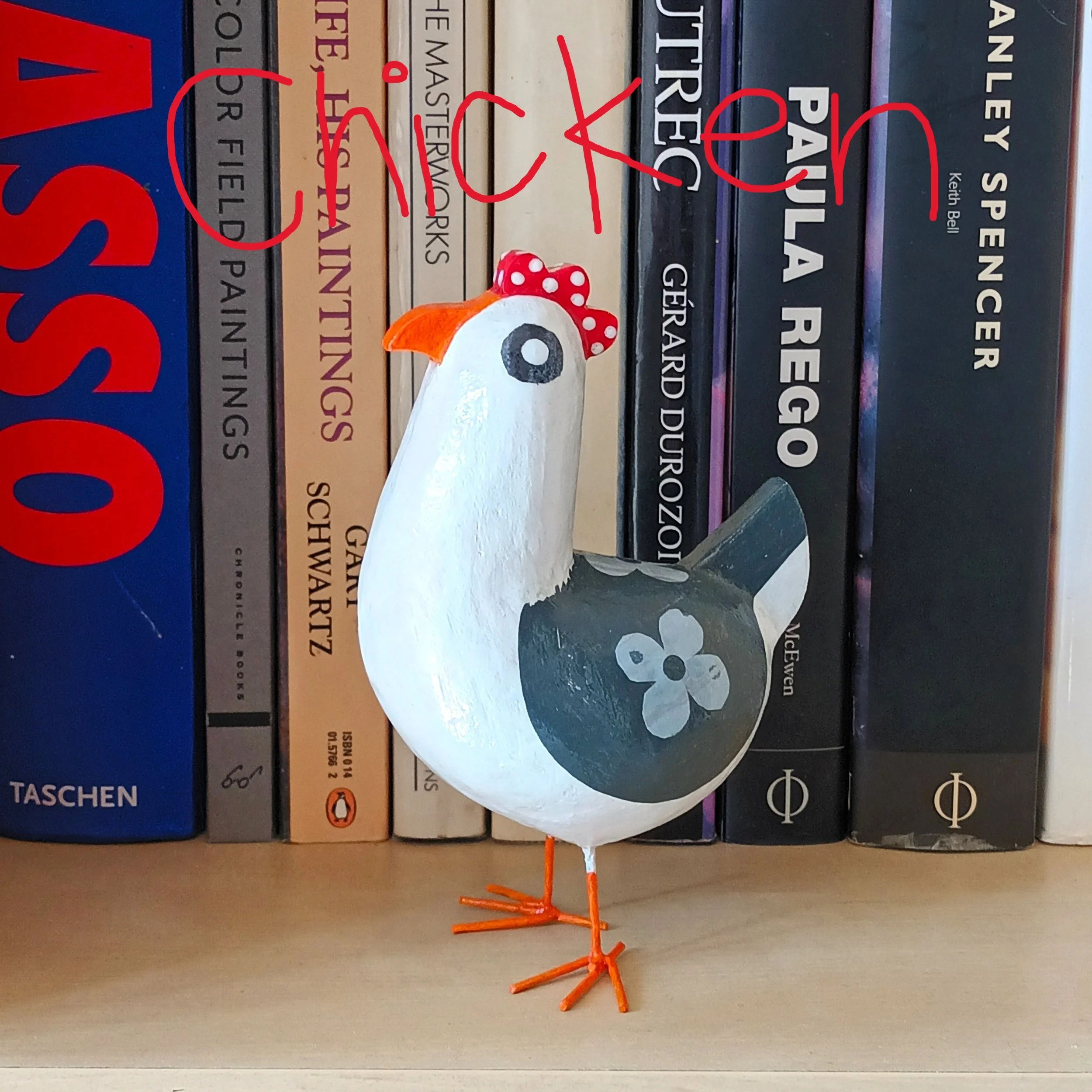 A small Posh Paper Mache mixed media chicken bird sculpture posed on a shelf in front of a selection of art books from the artists library, photographed in the artists home.
