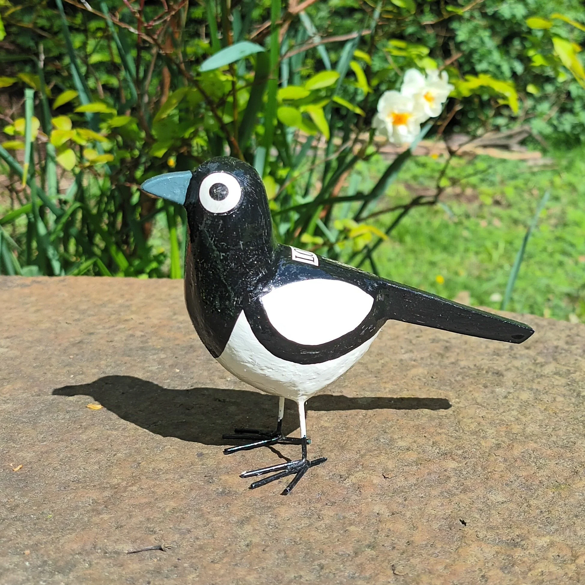 A small black and white decorative magpie bird posh paper mache sculpture with a grey beak, standing on a metal surface with garden plants and white flowers in the background photographed in the artists garden.