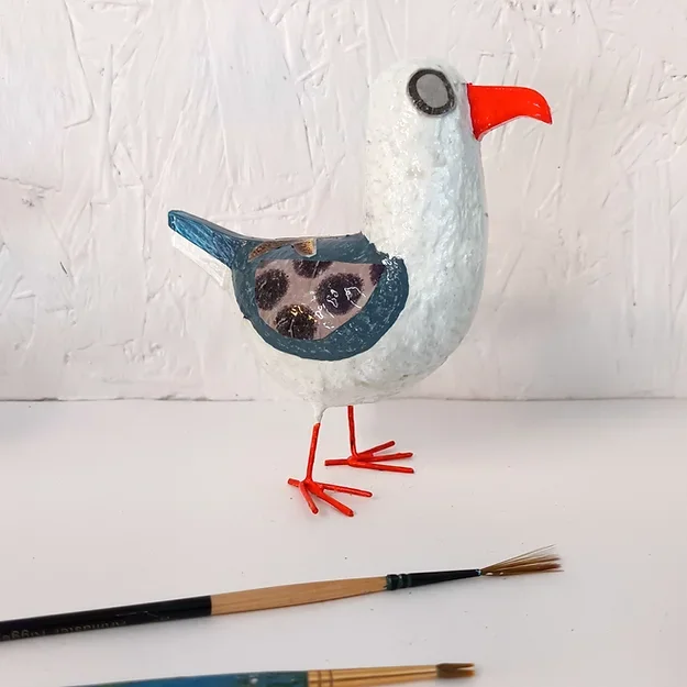 A decorative seagull posh paper mache sculpture by Tania Holland sculpture with a white body, black eye, red beak, and red legs, standing on a surface with paint brushes in front of it to give it scale.