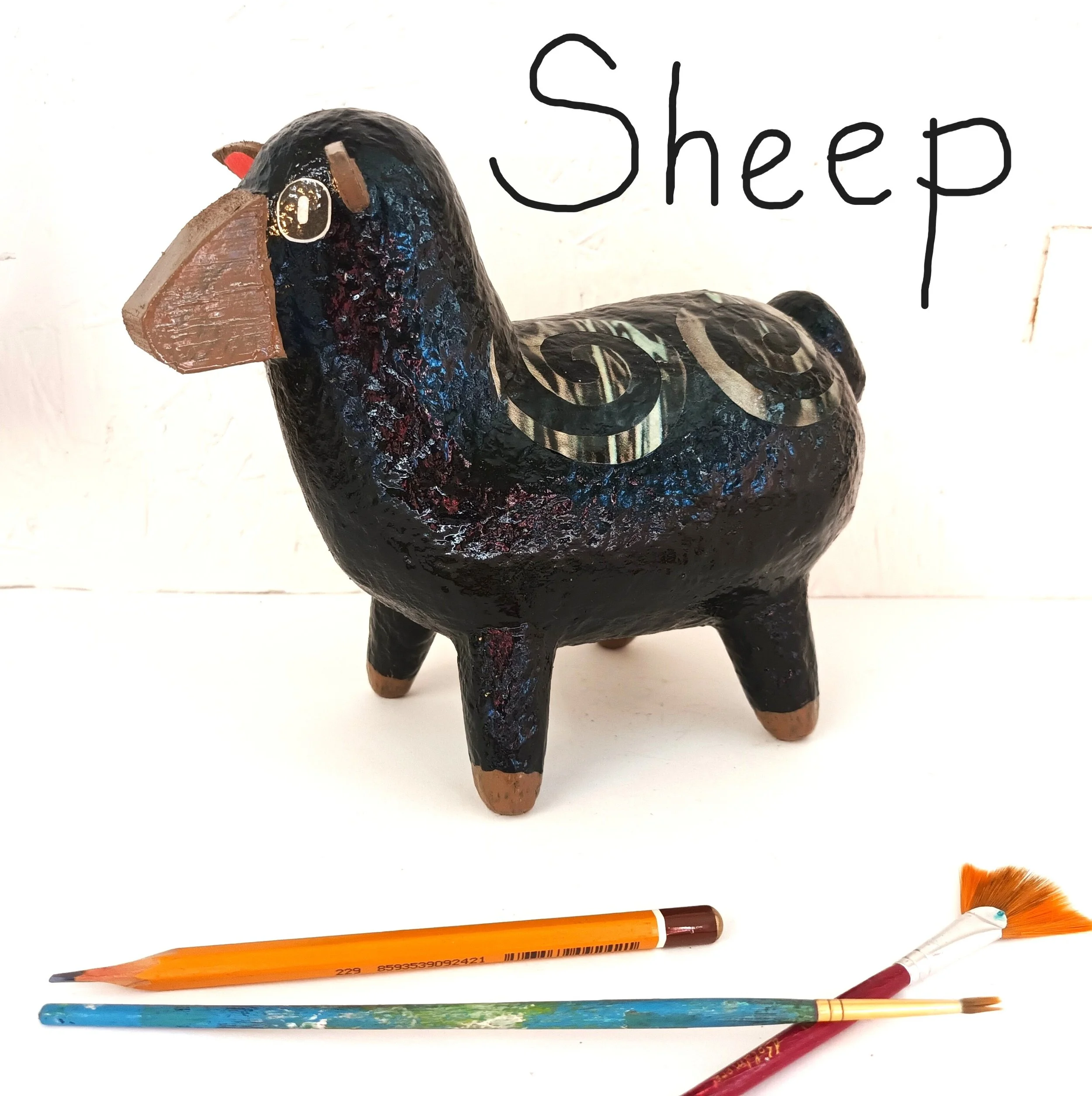 A medium sized Posh Paper Mache mixed media sheep standing next to a pencil and paint brushes for scale.  Photographed in the artists studio.