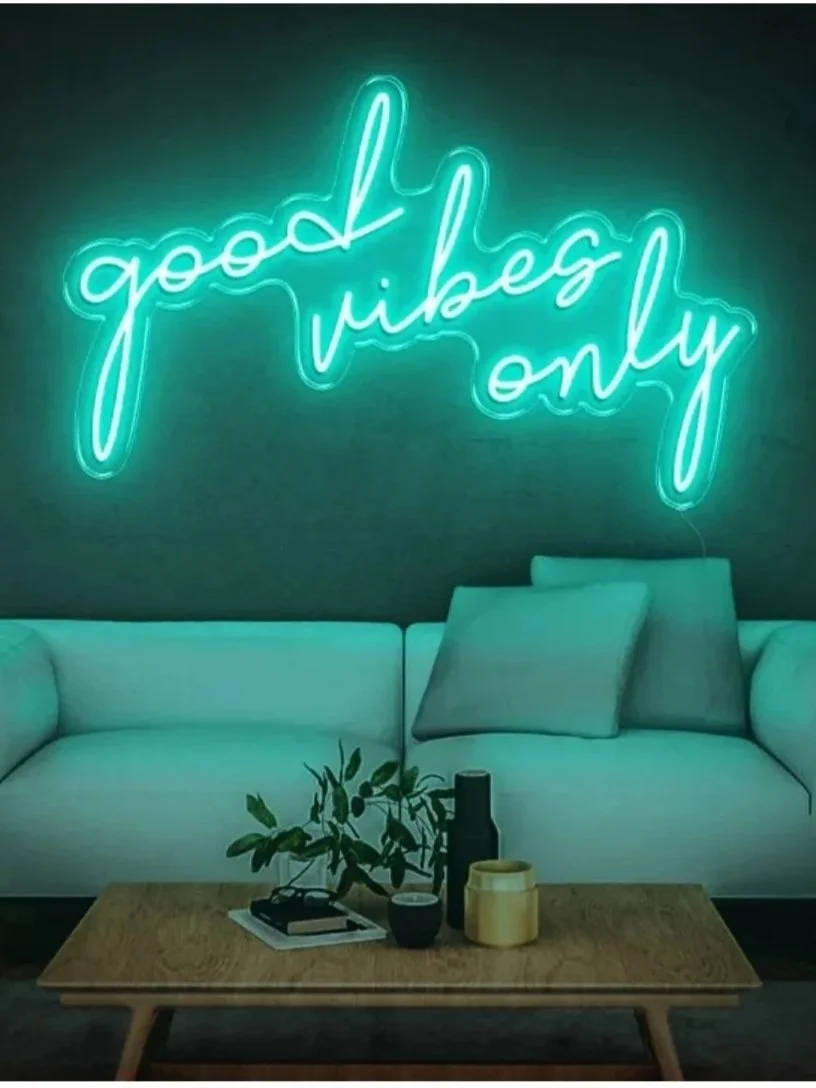 Neon sign reading 'good vibes only' hangs above a light-colored sofa with pillows in a modern living room.
