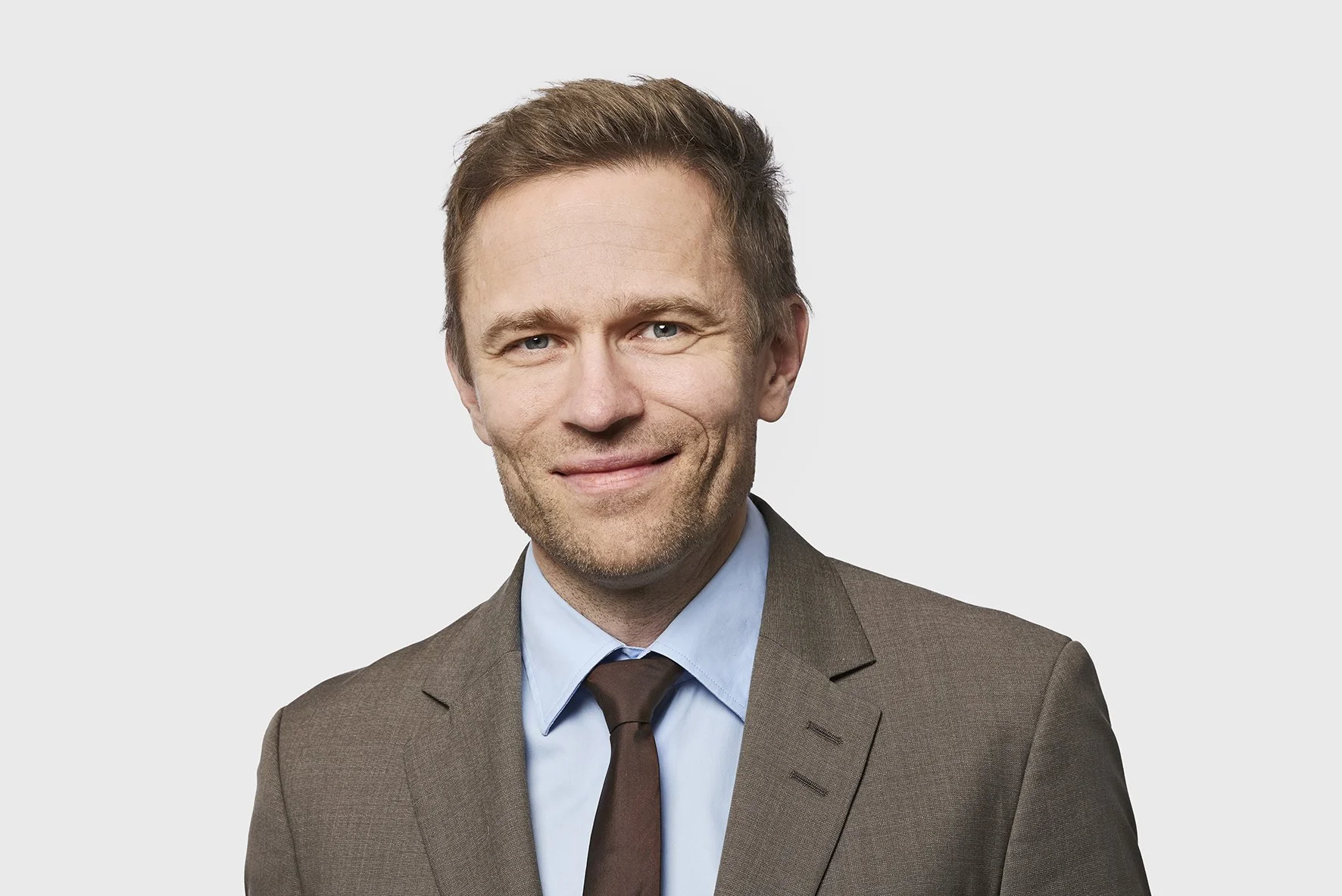 A man in a brown suit and blue shirt smiling.