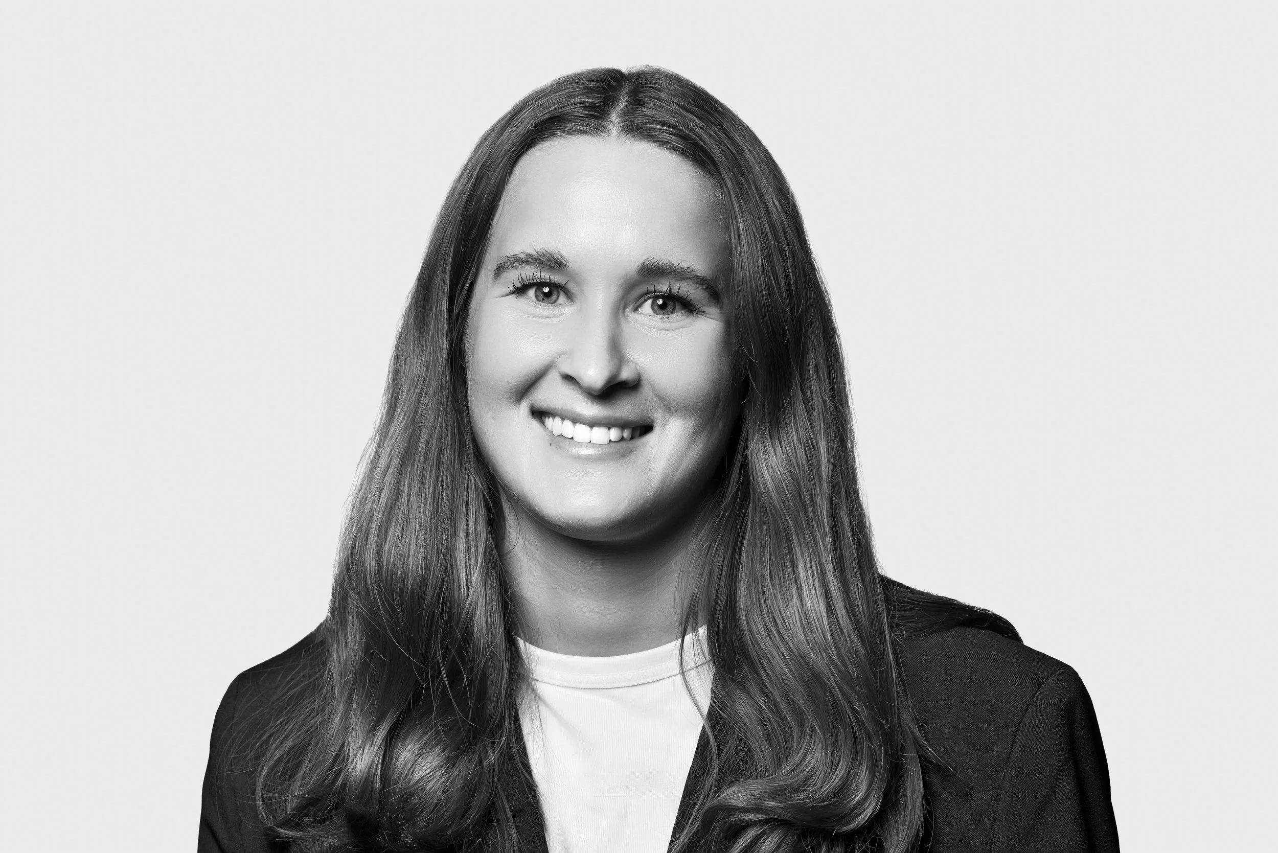 Black and white portrait of a young woman with long, wavy hair smiling, wearing a dark blazer and a white top against a plain background.