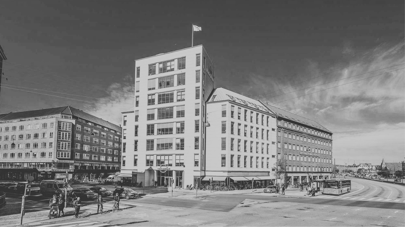 Black and white photo of a city street corner with modern and older buildings, parked cars, bicycles, and pedestrians at a crosswalk under a partly cloudy sky.