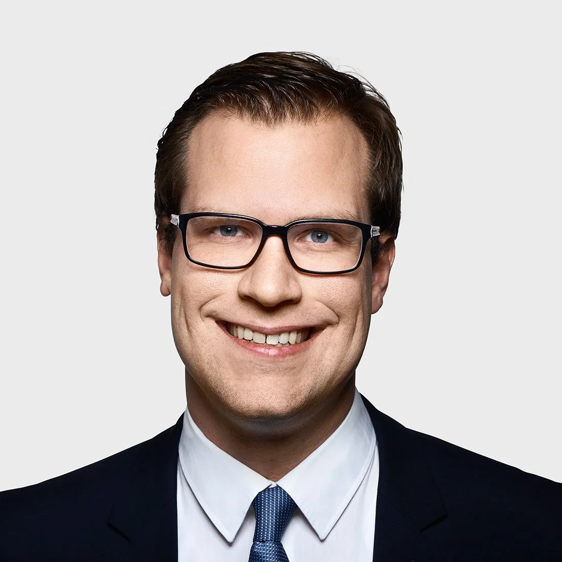 A professional headshot of a man with short brown hair, wearing black glasses, a dark suit, white shirt, and blue tie, smiling against a plain light grey background.