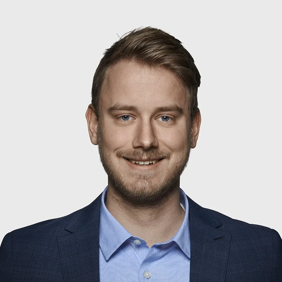 A portrait of a young man with light brown hair, blue eyes, and a light beard, smiling, wearing a navy blue blazer and a light blue dress shirt, against a plain light gray background.