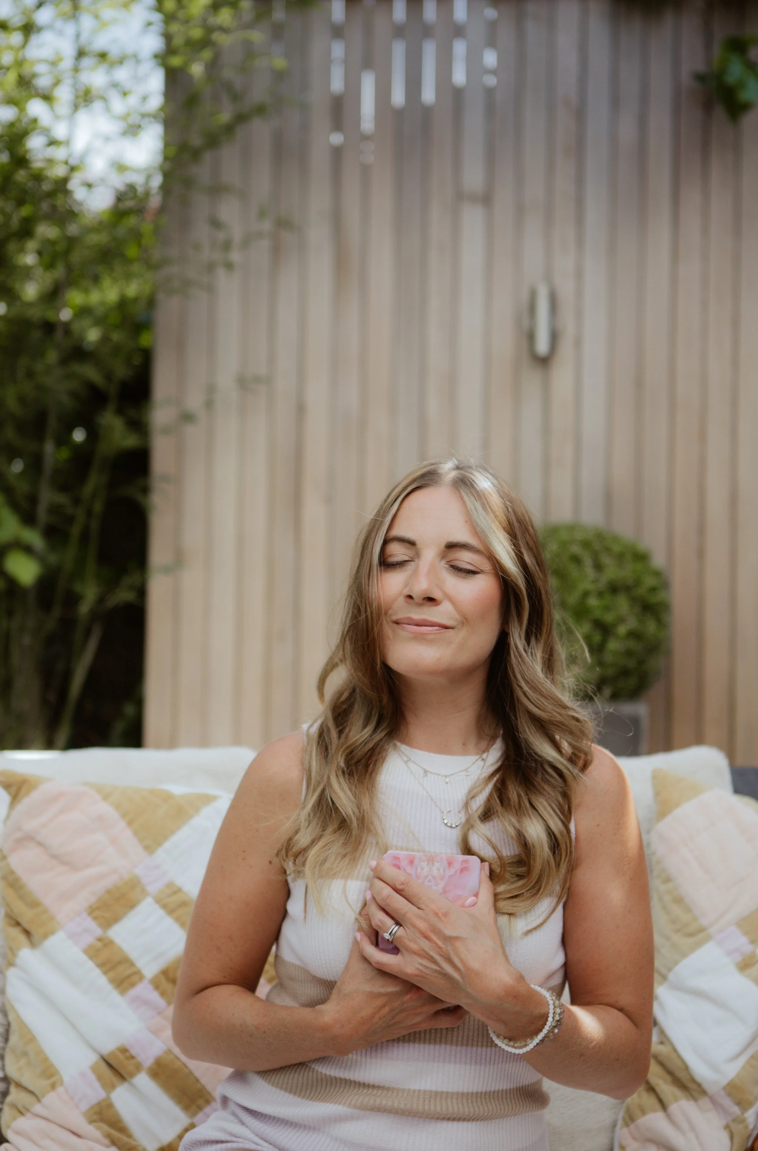 A woman sitting outdoors on a couch holding a pink phone with both hands, eyes closed, smiling, with a wooden fence and greenery in the background.