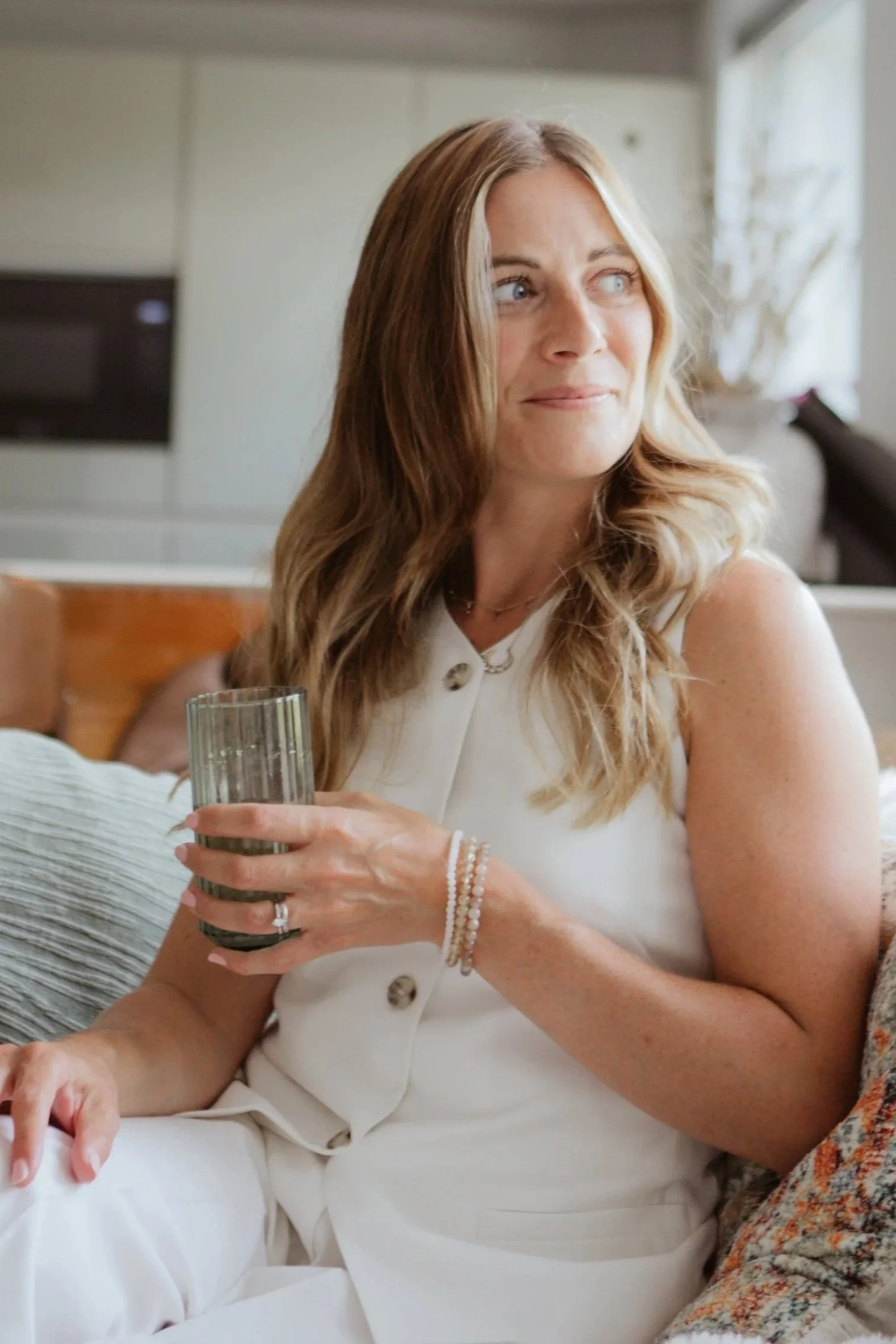 A woman with light brown, wavy hair sitting on a couch, holding a glass of water, wearing a white waist coat and matching trousers, accessorised with bracelets and a necklace, in a brightly lit room.
