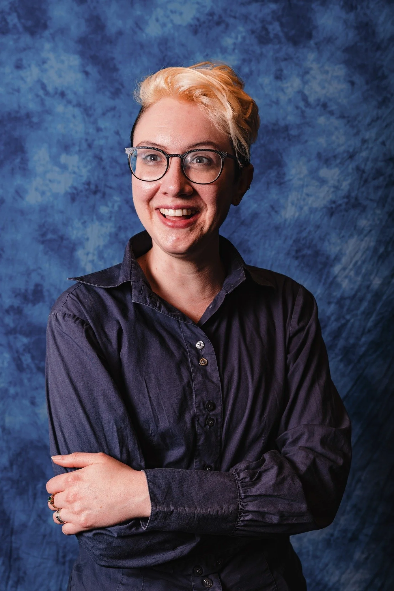 Woman with short, blonde hair, glasses, wearing a dark blue button-up shirt, smiling with arms crossed, posed against a blue textured background.