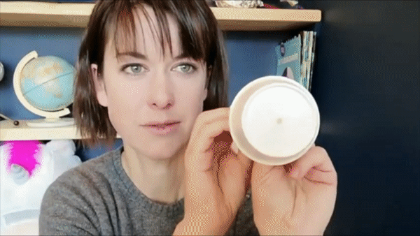 Person holding a white cup towards the camera in a room with a bookshelf and globe in the background.