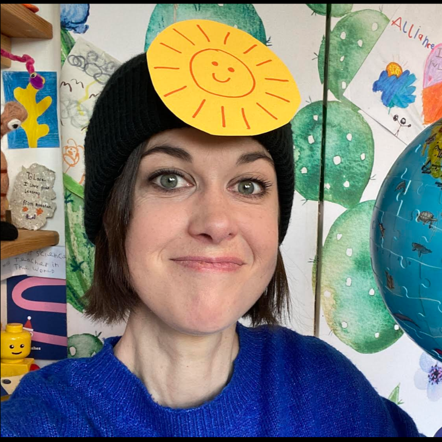Woman wearing a black knit hat with a paper sun on top, smiling in front of a colorful, childlike mural with cacti and drawings.