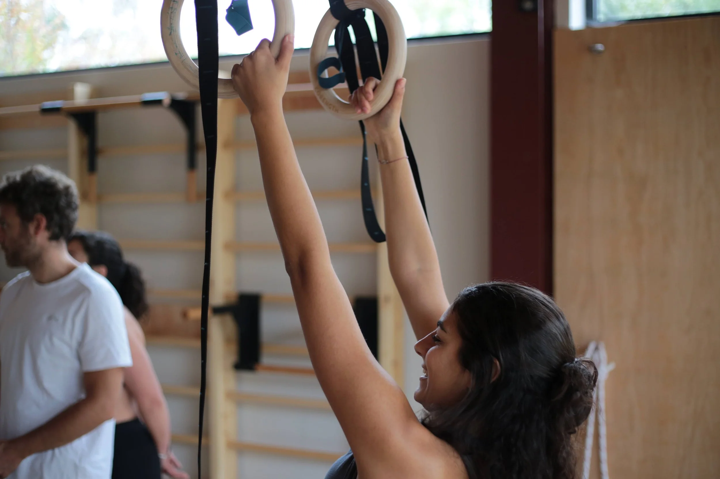 Woman using gymnastic rings in a fitness class, other people in the background, gym equipment and wooden wall bars visible.