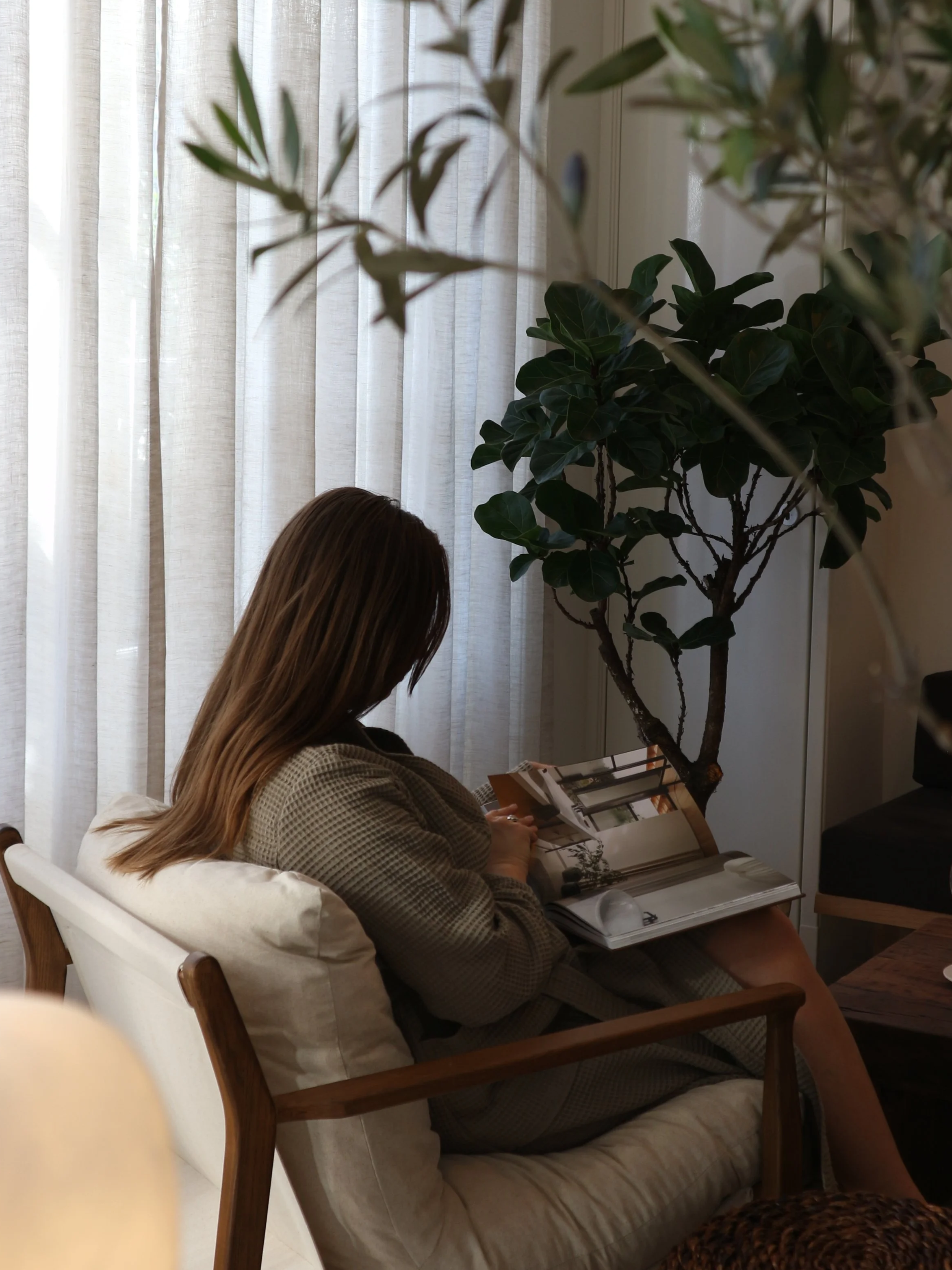A woman sitting on a cushioned chair reading a magazine, near a large potted plant and light-colored curtains.