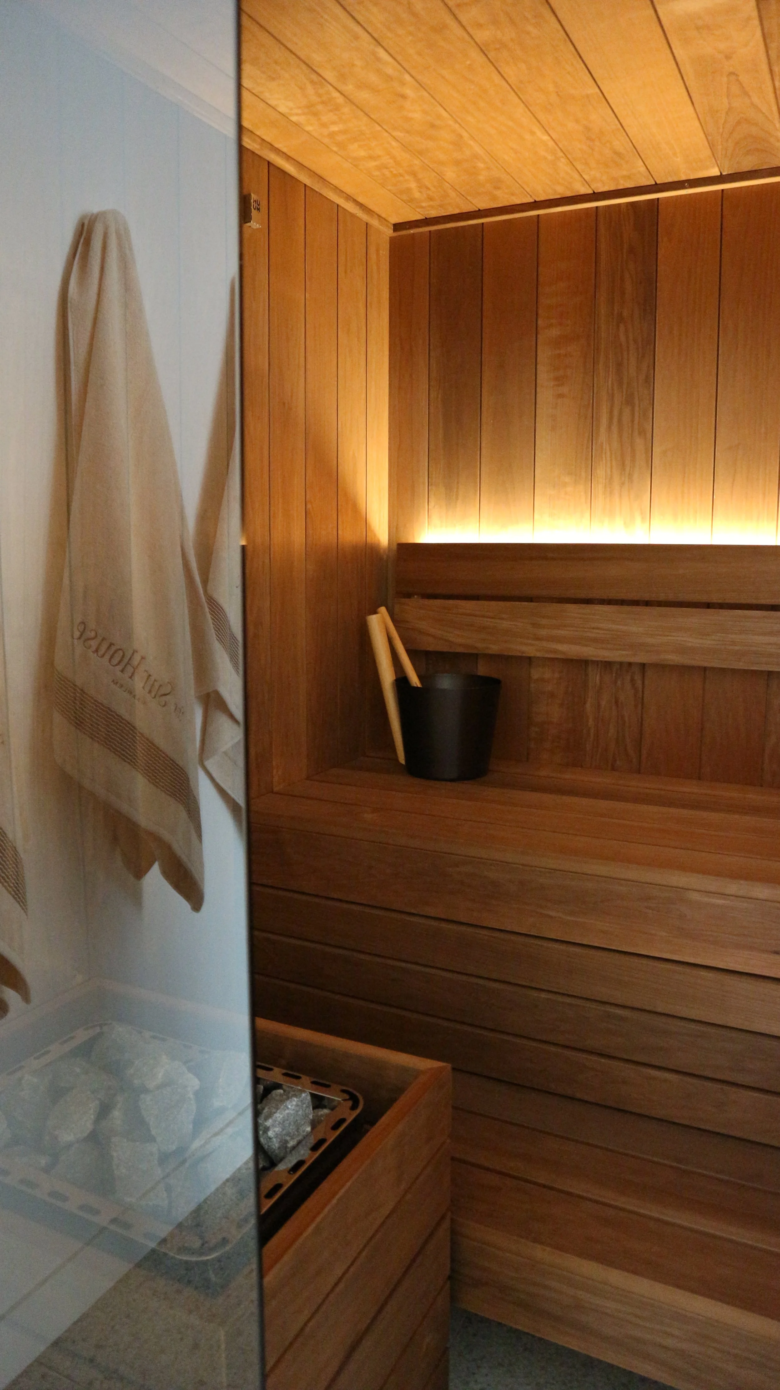 A sauna room with wooden paneling, a black container with a light-colored ladle and sauna brush, and a towel hanging on the wall.