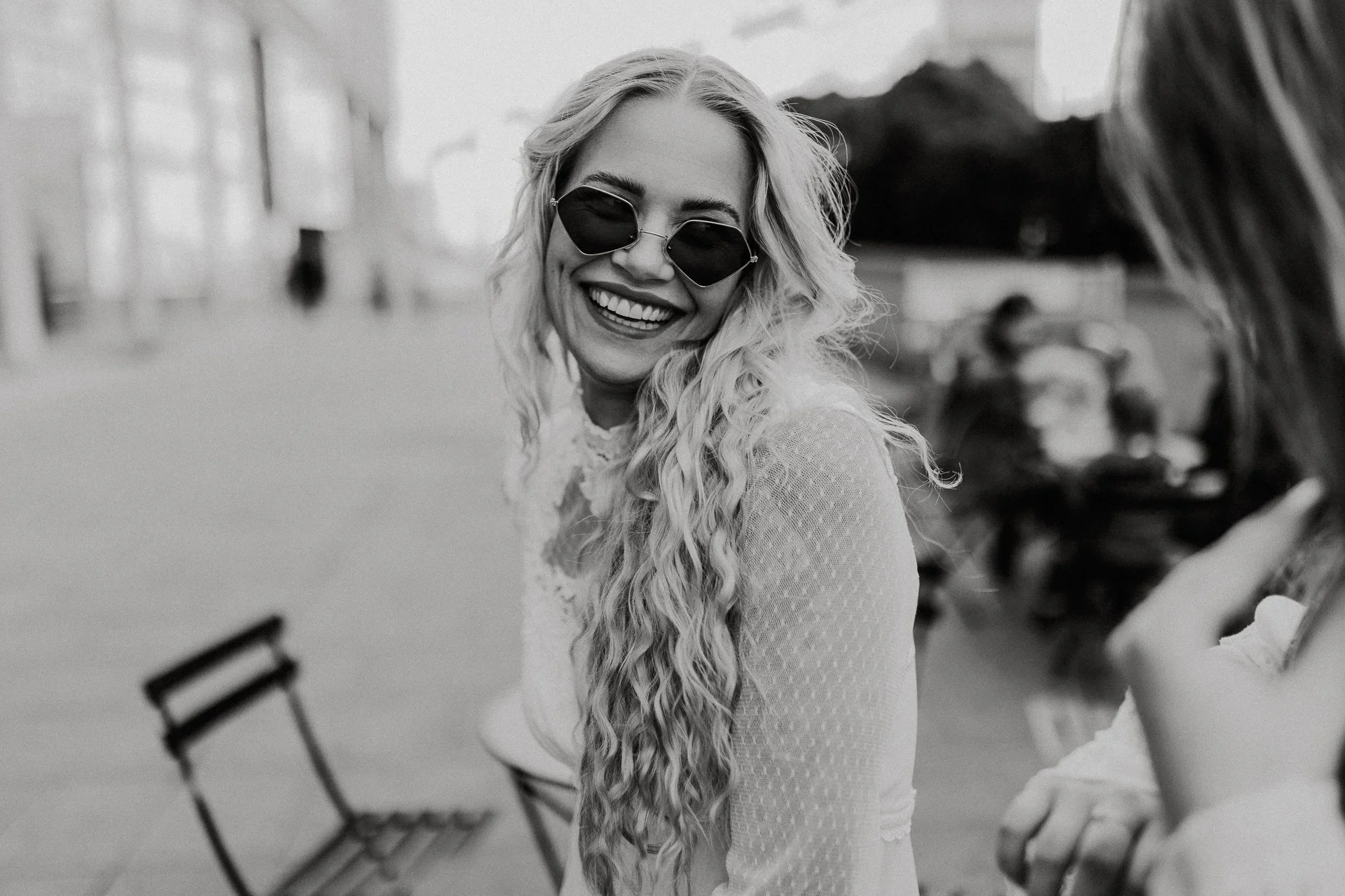 En kvinne med langt krøllete hår, solbriller og en lys, sbrudekjole smiler utendørs.