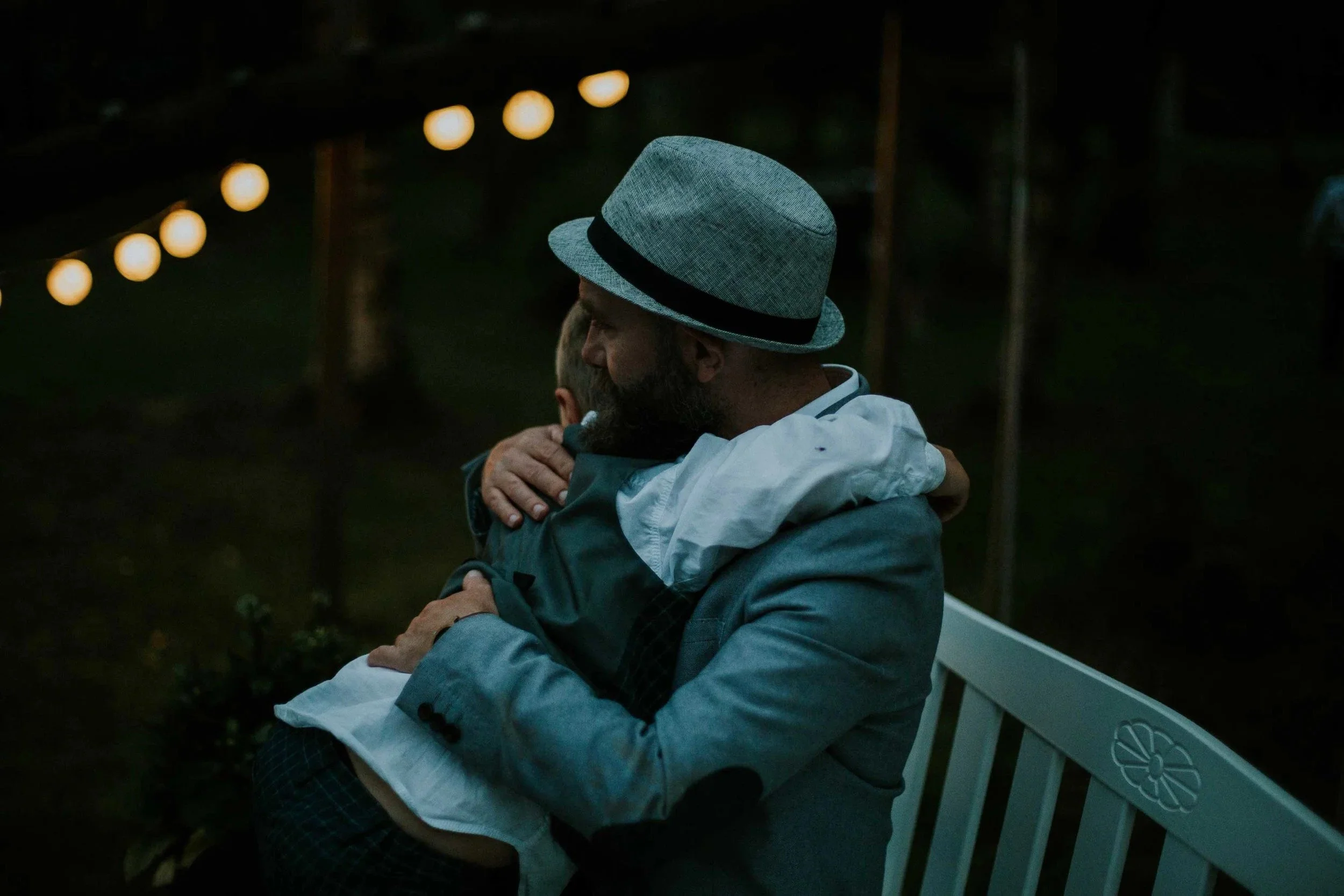 En mann med hatt og lys dressjakke klemmer en ung gutt på en hvit benk om kvelden. Runde lysslynger er uskarpe i bakgrunnen.