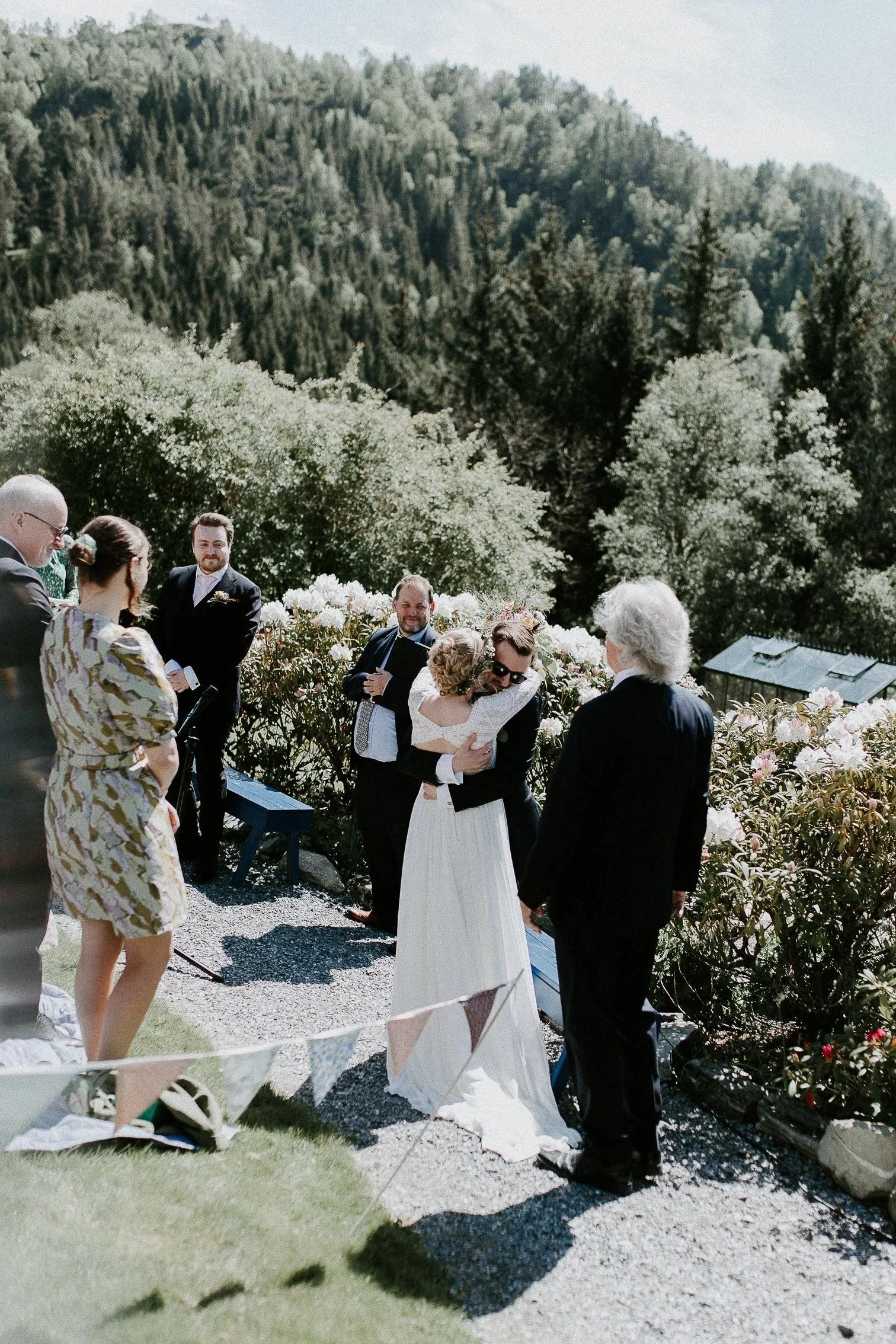 Utendørs bryllupsseremoni med fjell og grønn natur i bakgrunnen. Brud og brudgom omfavner hverandre omgitt av gjester.
