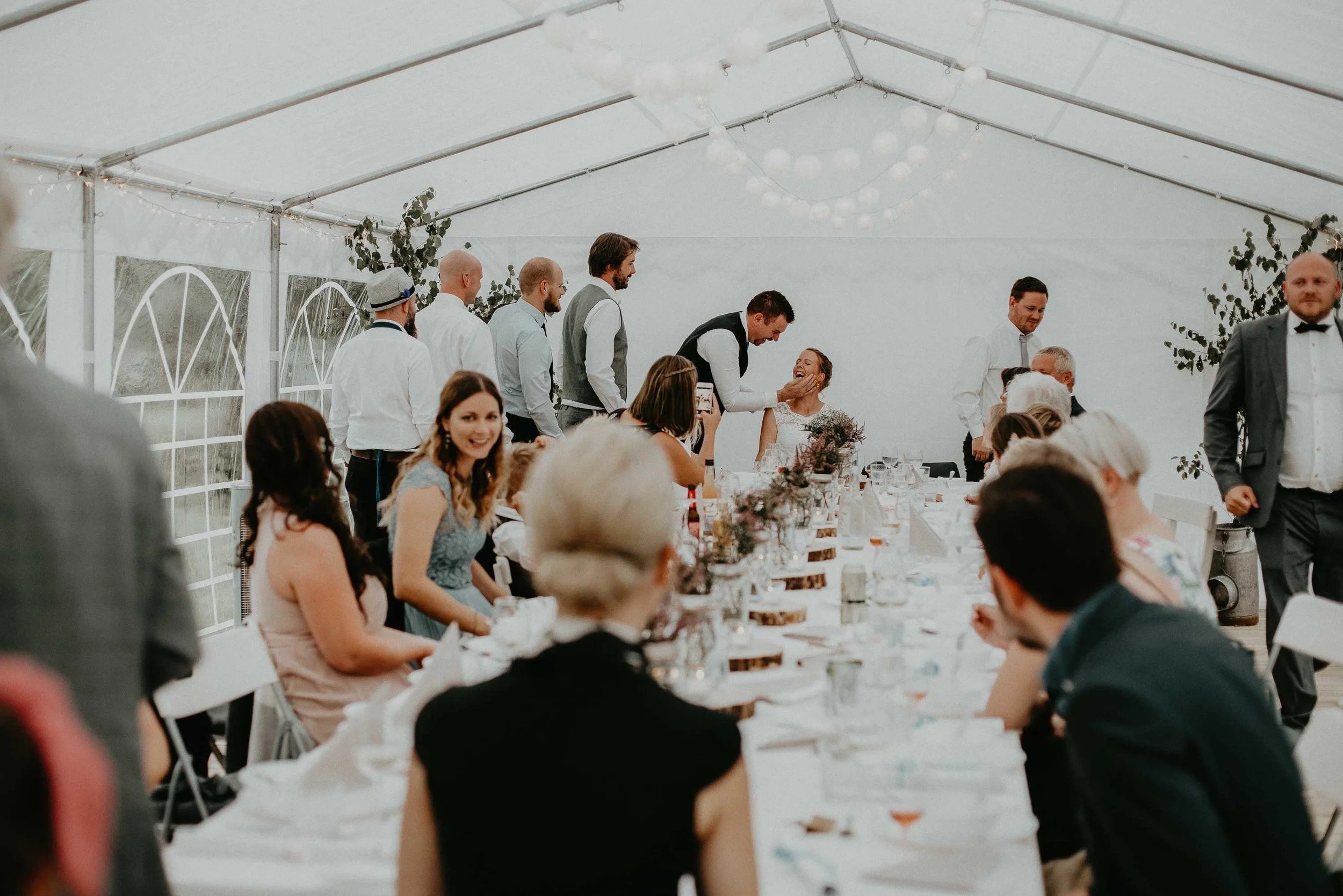 Bryllupsfest i et stort hvitt telt med langbord. Noen gjester sitter, andre står, mens brudeparet er samlet ved enden av bordet.