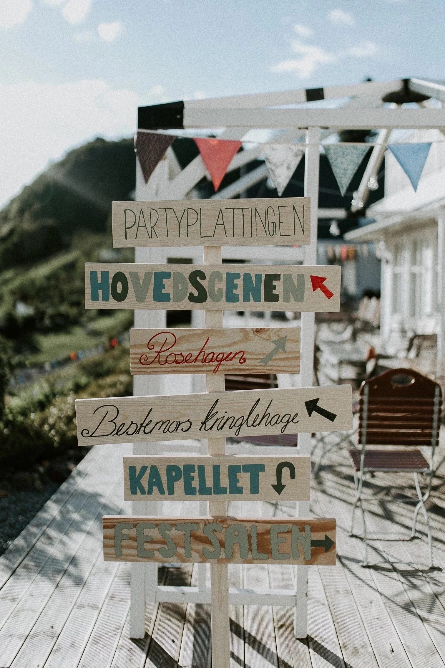 Dekorativ retningsskilt med flere piler som peker i ulike retninger, plassert utendørs på en treplatting med stoler og vimpler.