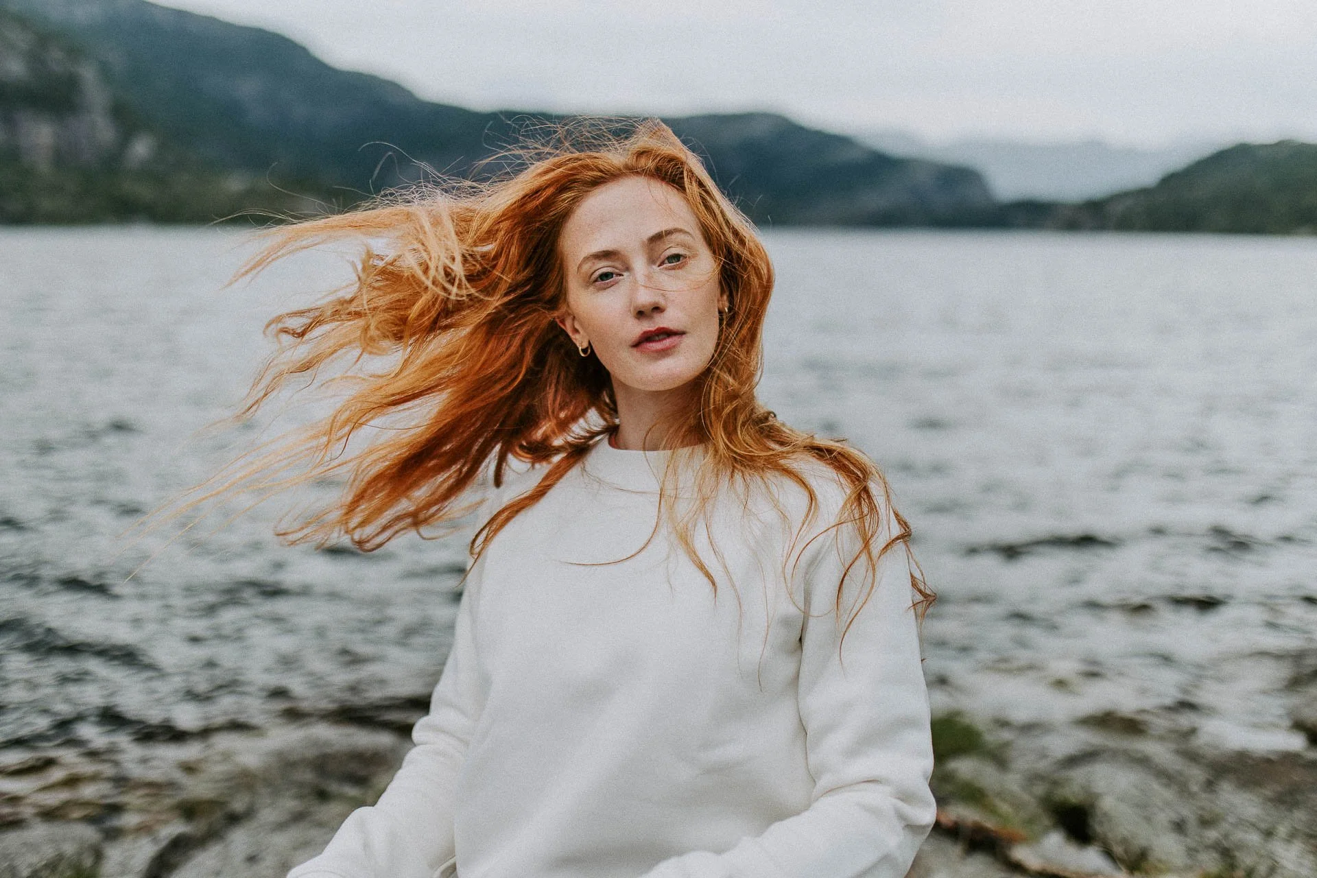 Kvinne med rødt hår i vind ser i kameraet fjell og vann i bakrunnen hvit genserReklamefotografering til kampanje for Skogstad bilder tatt i preikestolenområdet