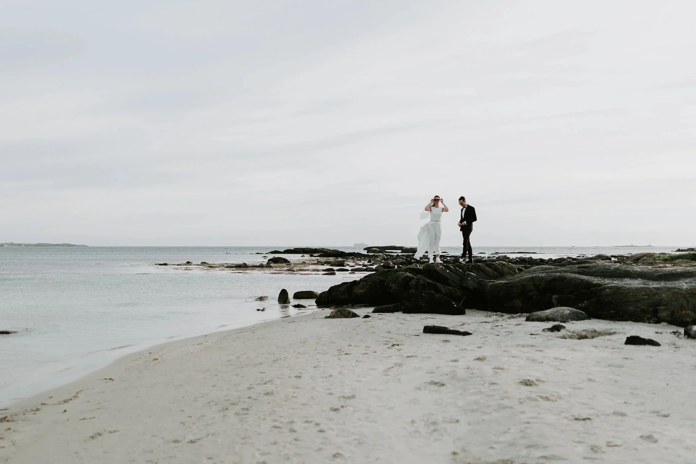 Et par i bryllupsantrekk står på steiner ved stranden. Den ene justerer solbriller mens den andre ser ned. Hav og overskyet himmel i bakgrunnen.