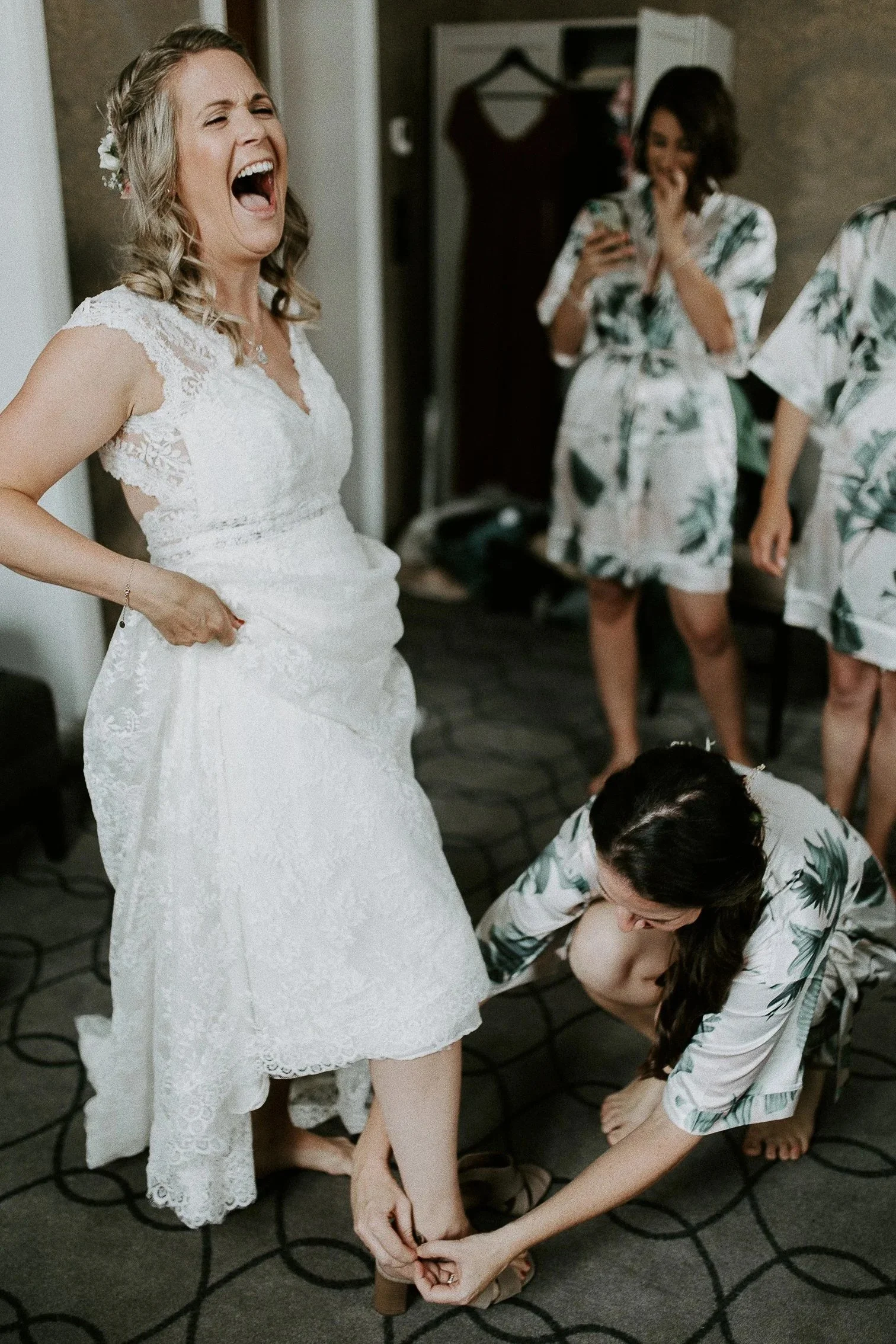 En brud i hvit blondekjole ler mens hun får hjelp til å ta på skoene. Forlovere i matchende morgenkåper med tropisk mønster i bakgrunnen.