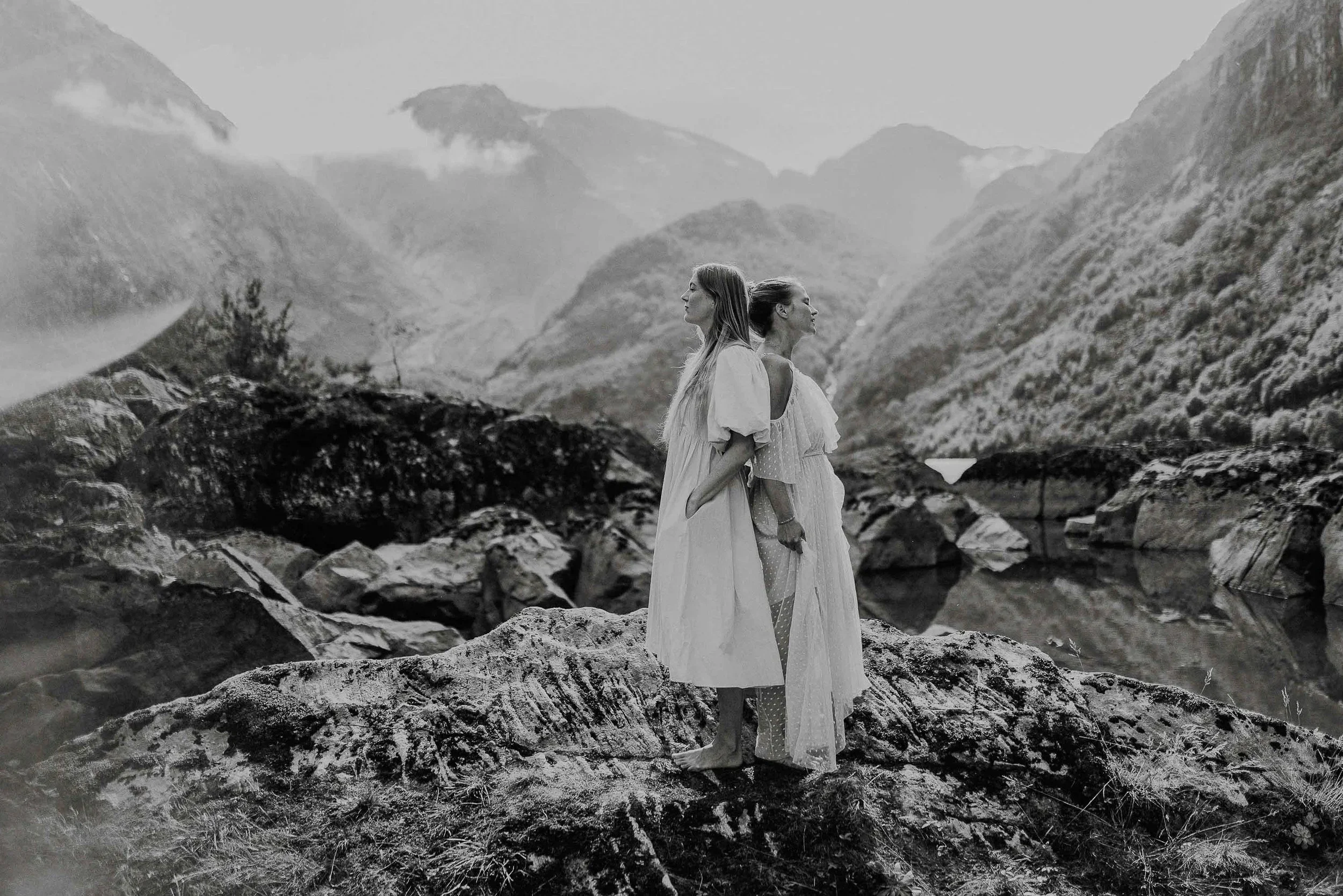 To kvinner står på steiner ved en elv i et fjellandskap, vendt bort fra hverandre. Sort hvitt fotografi.