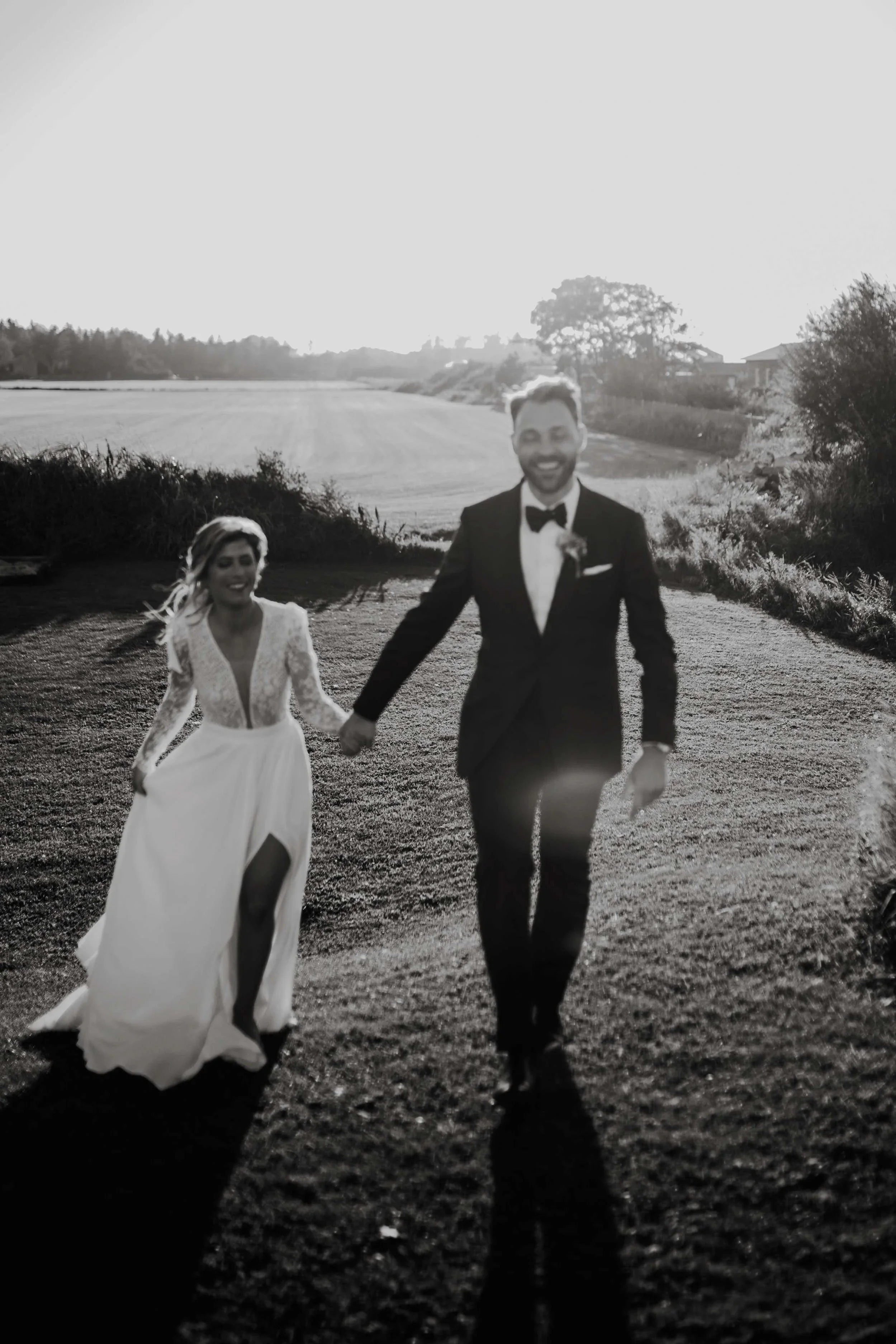 Sort hvitt bilde av et nygift par som går hånd i hånd utendørs på en grusvei sent på dagen, med jorde og trær i bakgrunnen.