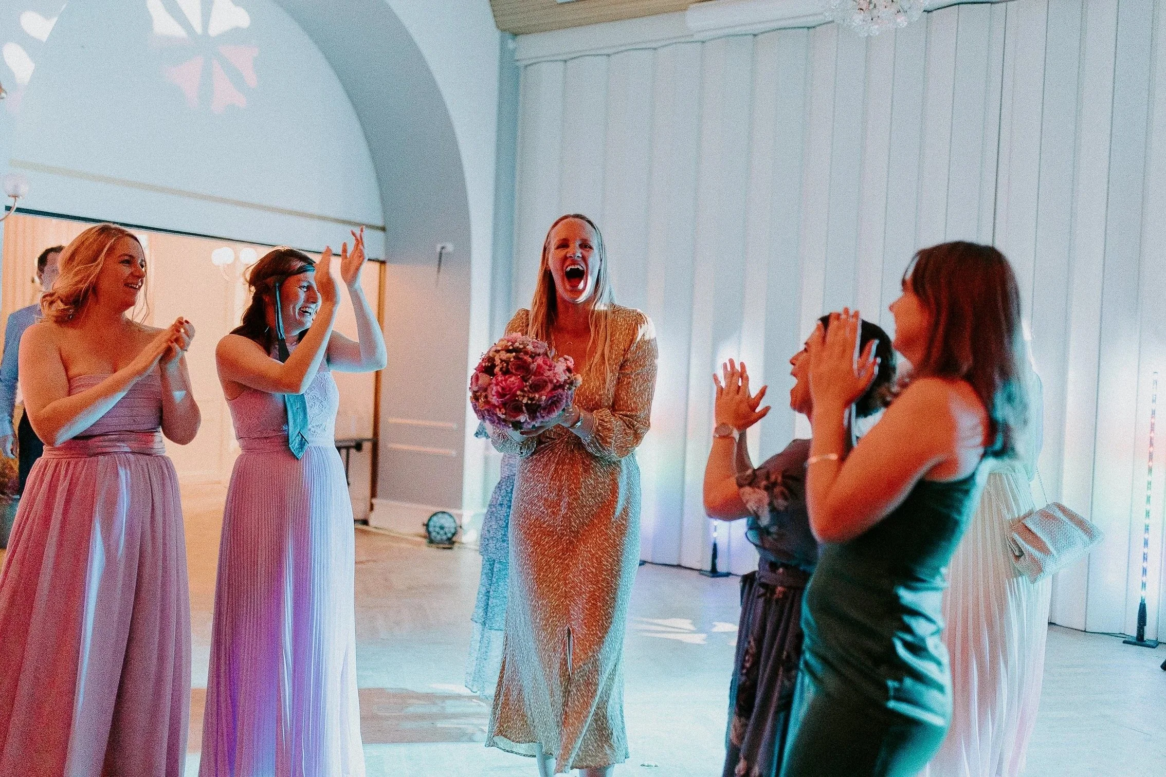 Brud, venner og gjester feirer under bryllupsfesten. En kvinne med bukett ler mens andre klapper og deltar.