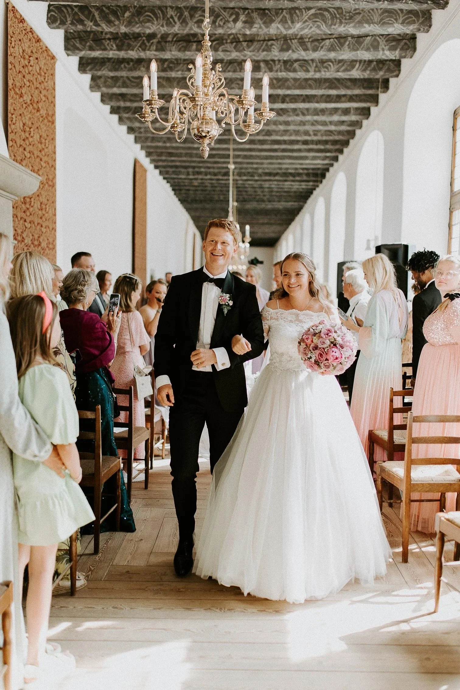 Brud og brudgom går ned midtgangen under bryllupsseremonien. Bruden holder en bukett med rosa roser. Gjestene sitter på begge sider og noen tar bilder. Seremonien er innendørs med store vinduer, lysekrone og tretak.