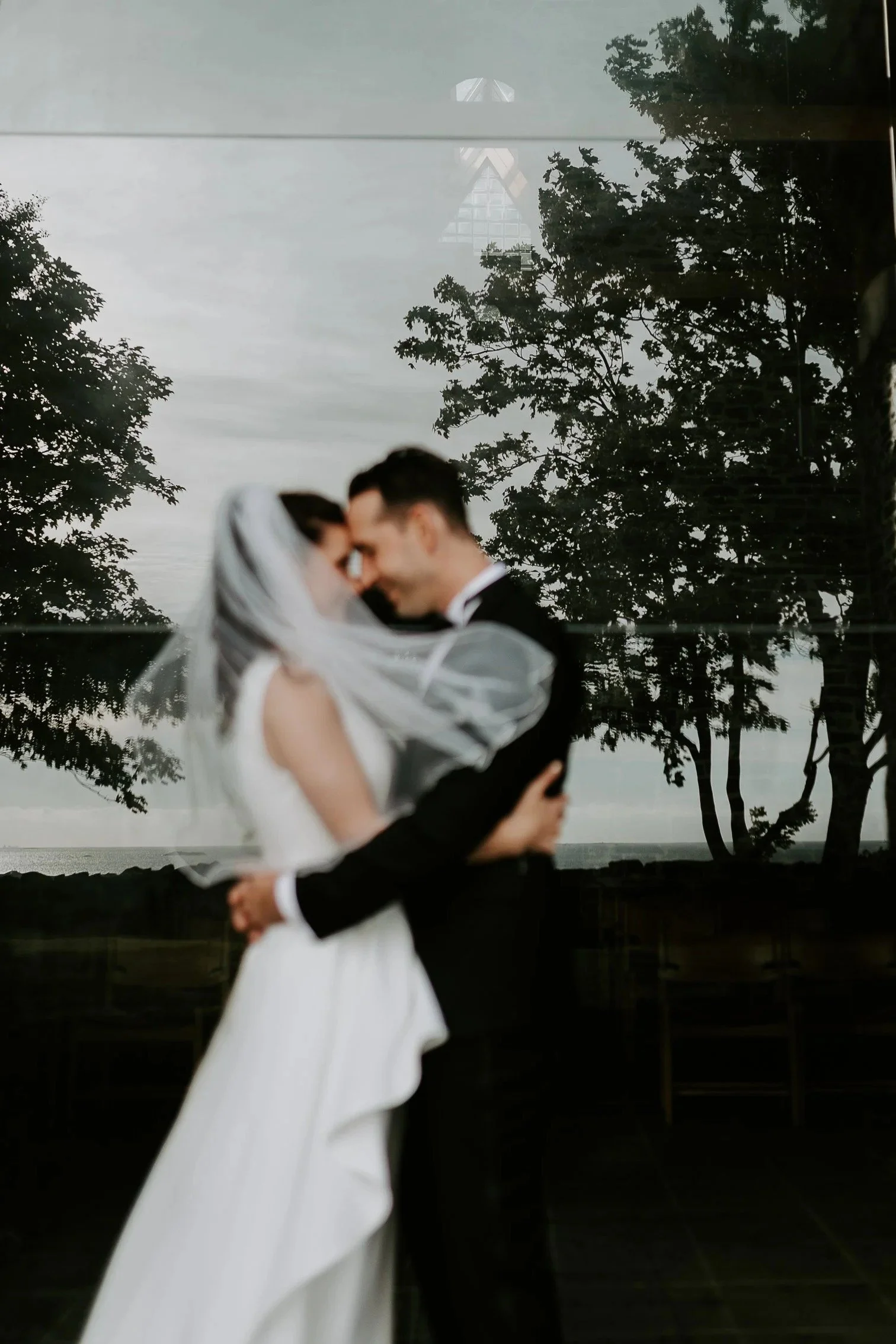 Uskarpt bilde av brud og brudgom som omfavner hverandre og berører pannen utendørs nær vann, med trær i bakgrunnen.