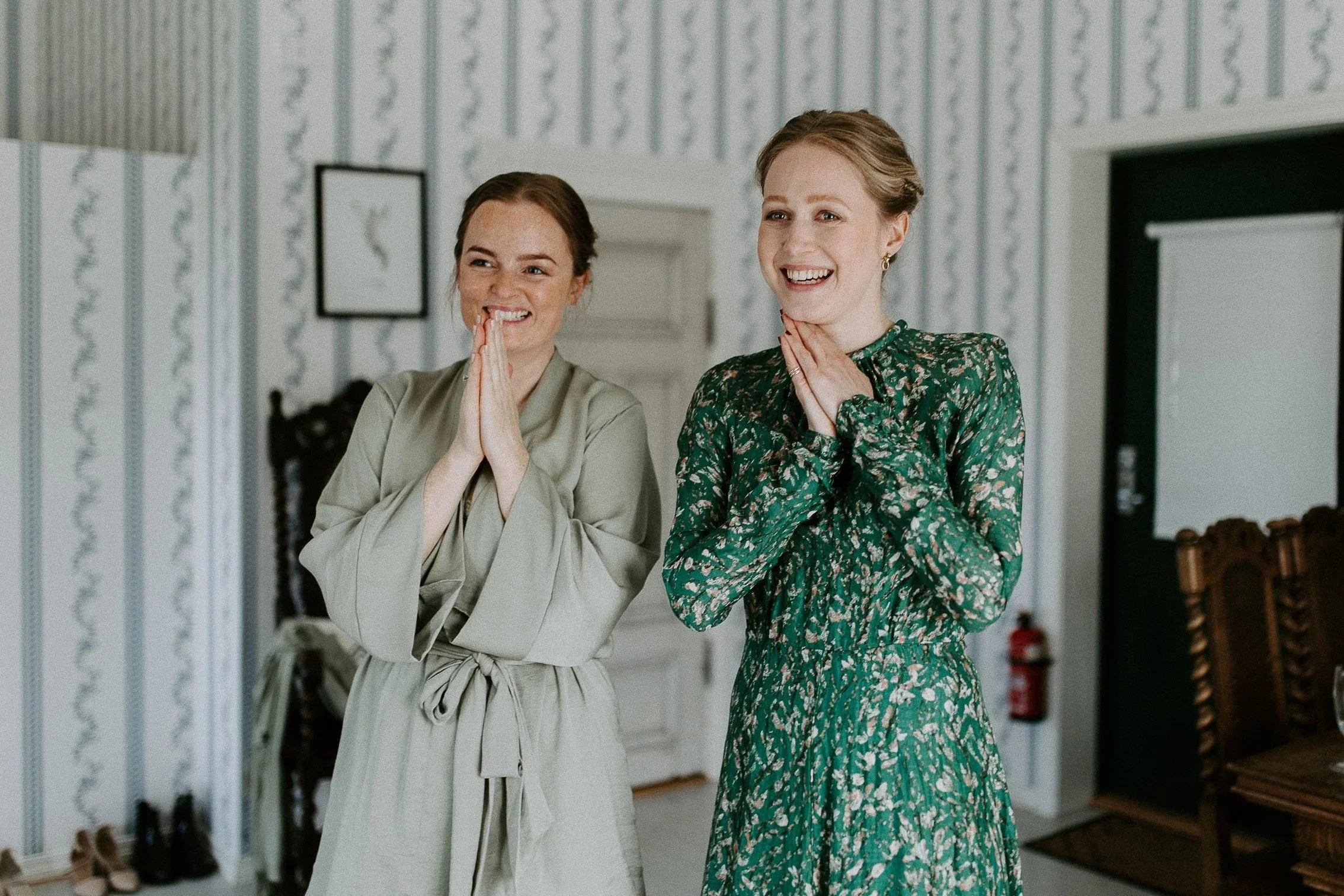 To kvinner står innendørs, smiler og klapper, med mønstret tapet og tremøbler i bakgrunnen.