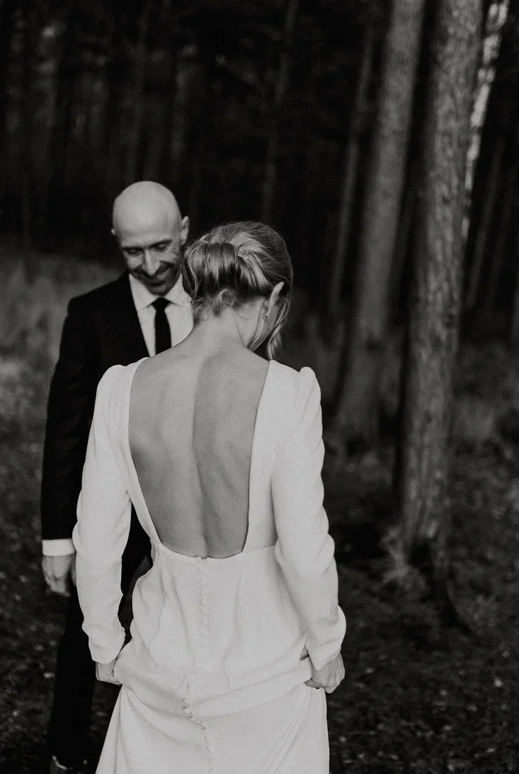 Et sort hvitt bilde av en kvinne i en ryggløs hvit kjole som står i skogsområde, med en mann i sort dress og slips smilende bak henne.