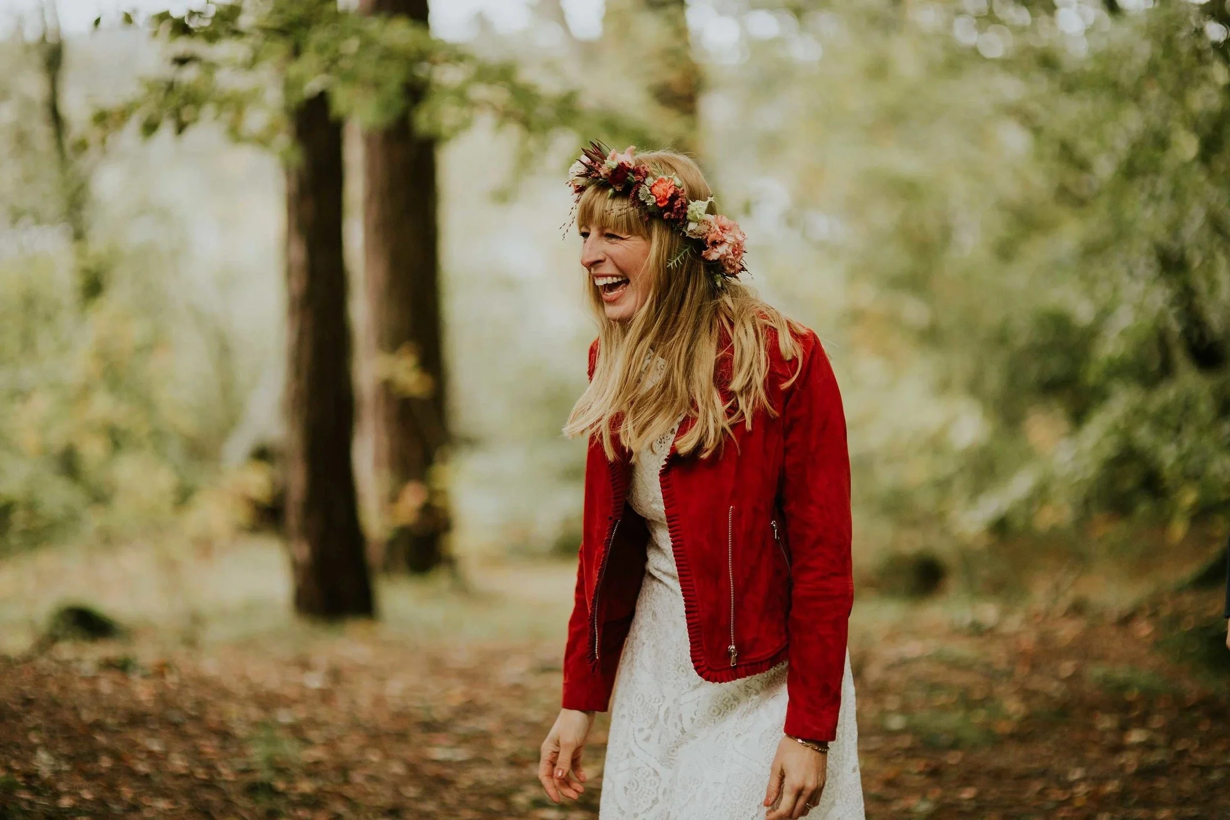 En kvinne med blomsterkrans, rød jakke og hvit kjole smiler og ler utendørs i skogsområde.