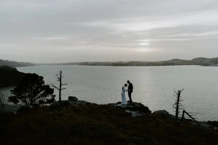 Et par står på en steinete høyde ved en innsjø, med trær og overskyet himmel i bakgrunnen i solnedgang.
