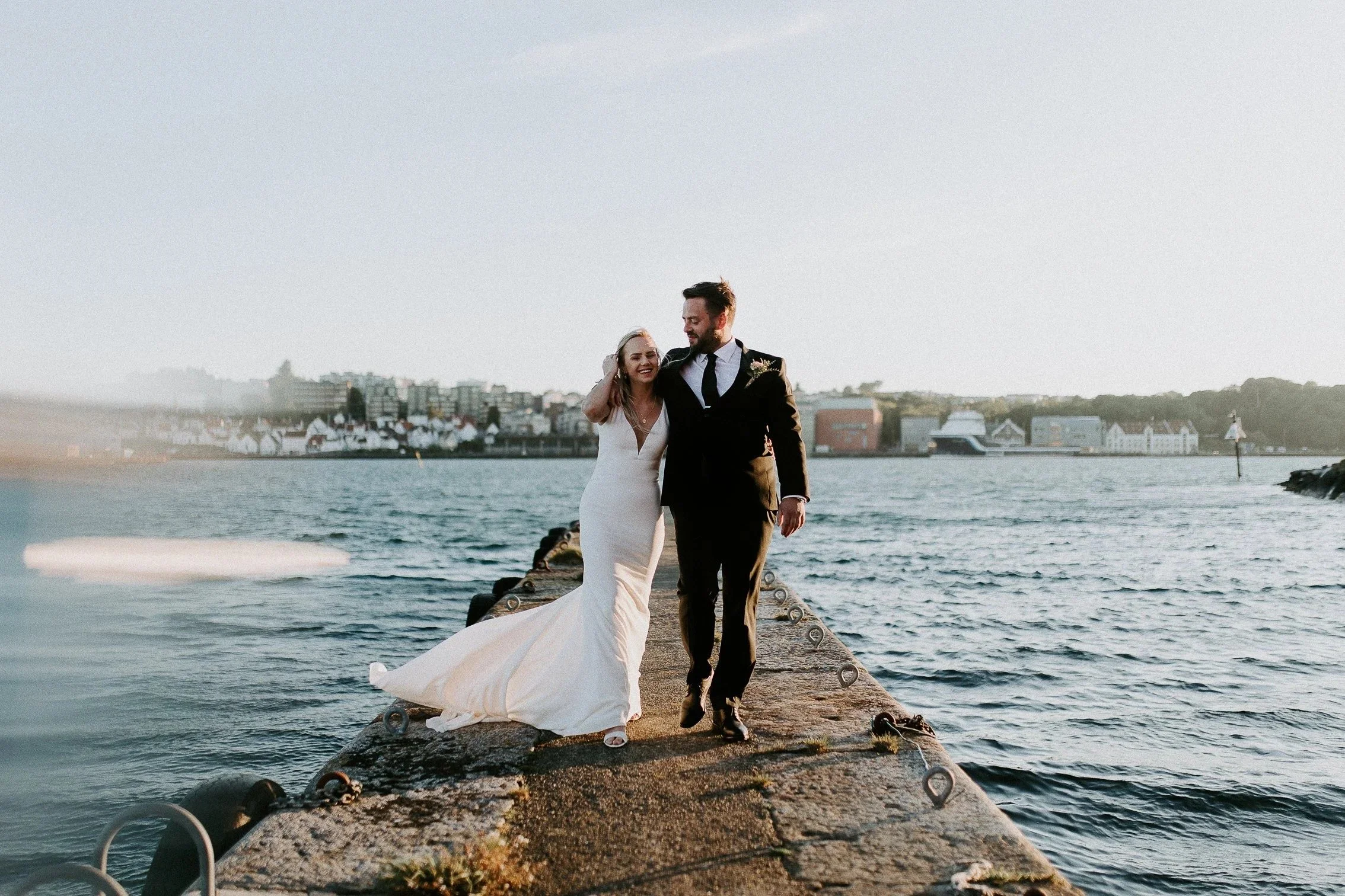 Et brudepar går sammen på en brygge ved vannet, smiler og holder hender, med en by i bakgrunnen.