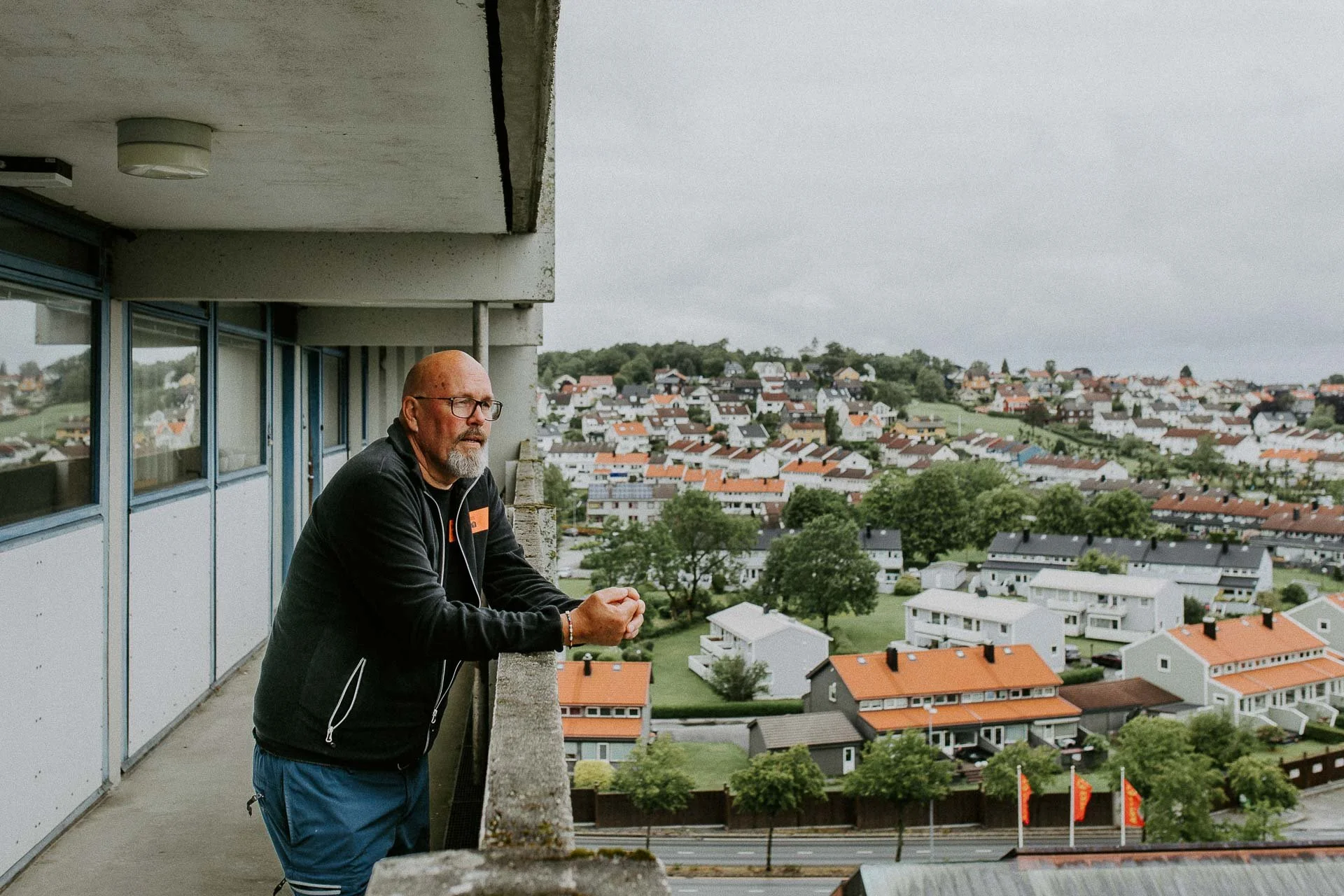 Mannen som som står og ser utover fra balkong høyt oppe i høyblokk i Hillevåg han ser rolig utover holder hender på rekkverk har briller naturlig lys, reklamefotografering for stavanger kommune, områdesatsingen i hillevåg