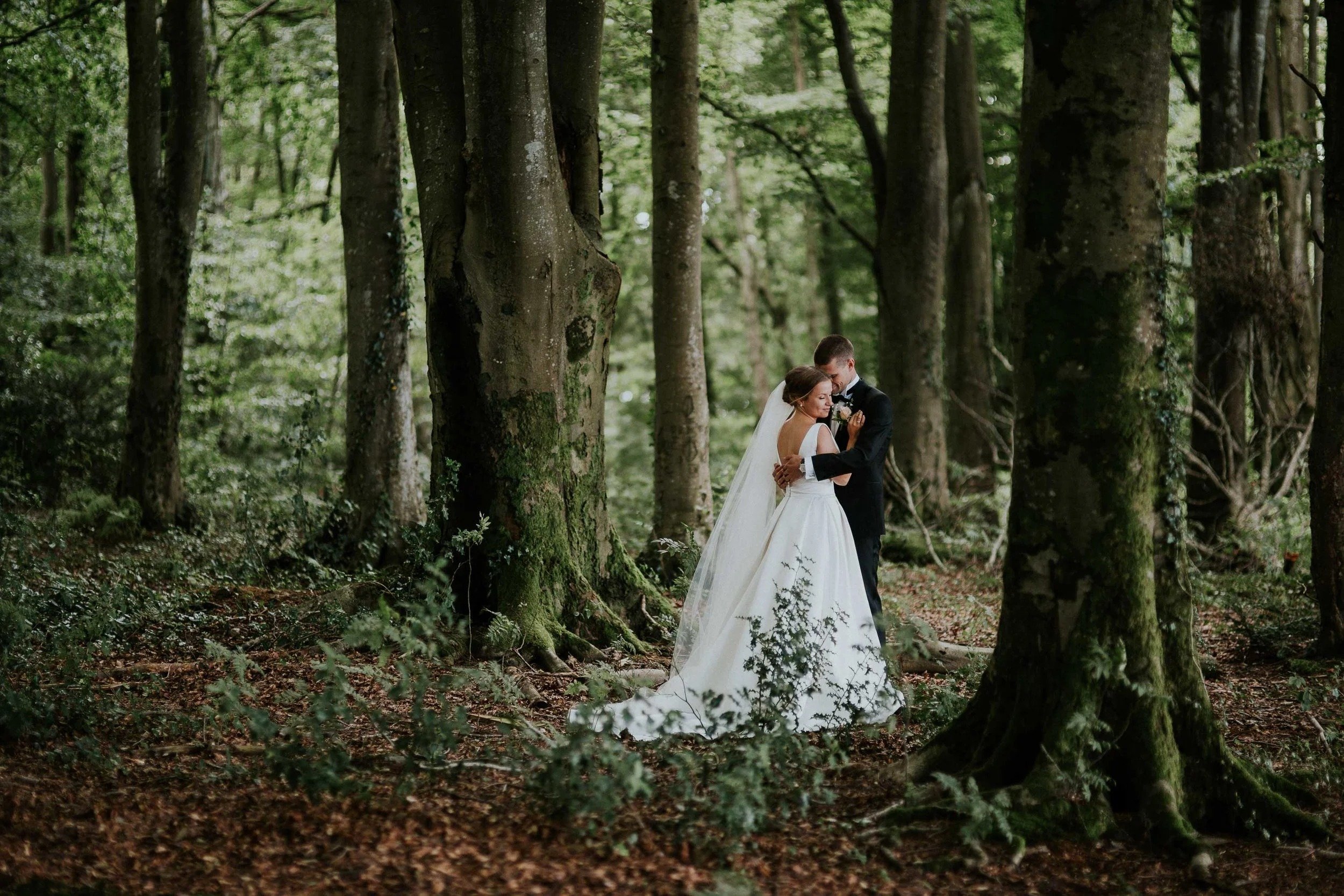 Brud og brudgom står tett sammen i skogen og deler et stille øyeblikk, omgitt av høye trær.