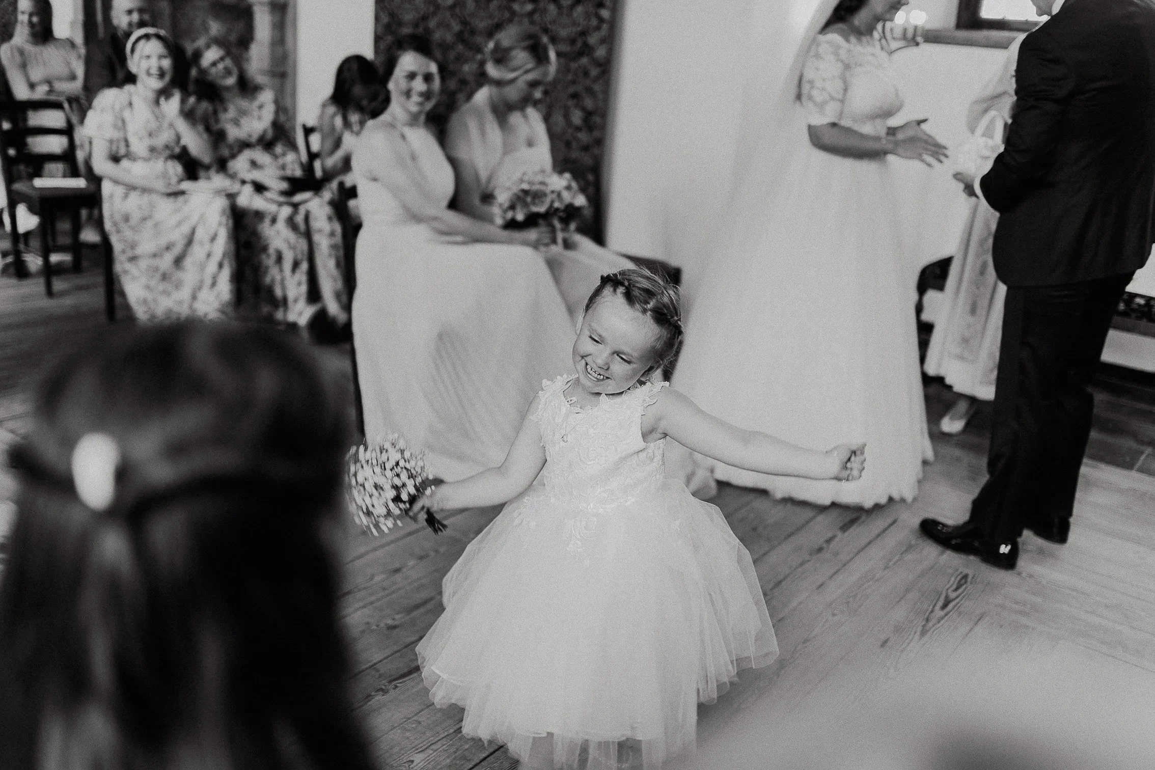 En brudepike i hvit kjole danser og smiler under bryllupsfesten, med gjester sittende i bakgrunnen.