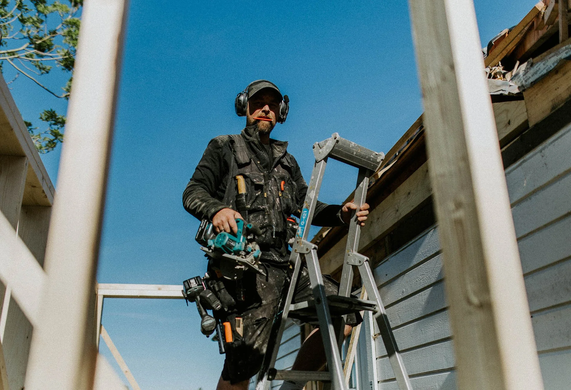 Reklamefotografering til kampanje for Altibox bedrift tatt i naturlig lys bilde av tømrer som er i arbeidssituasjon pusser opp hus står i stige med bylant i munnen