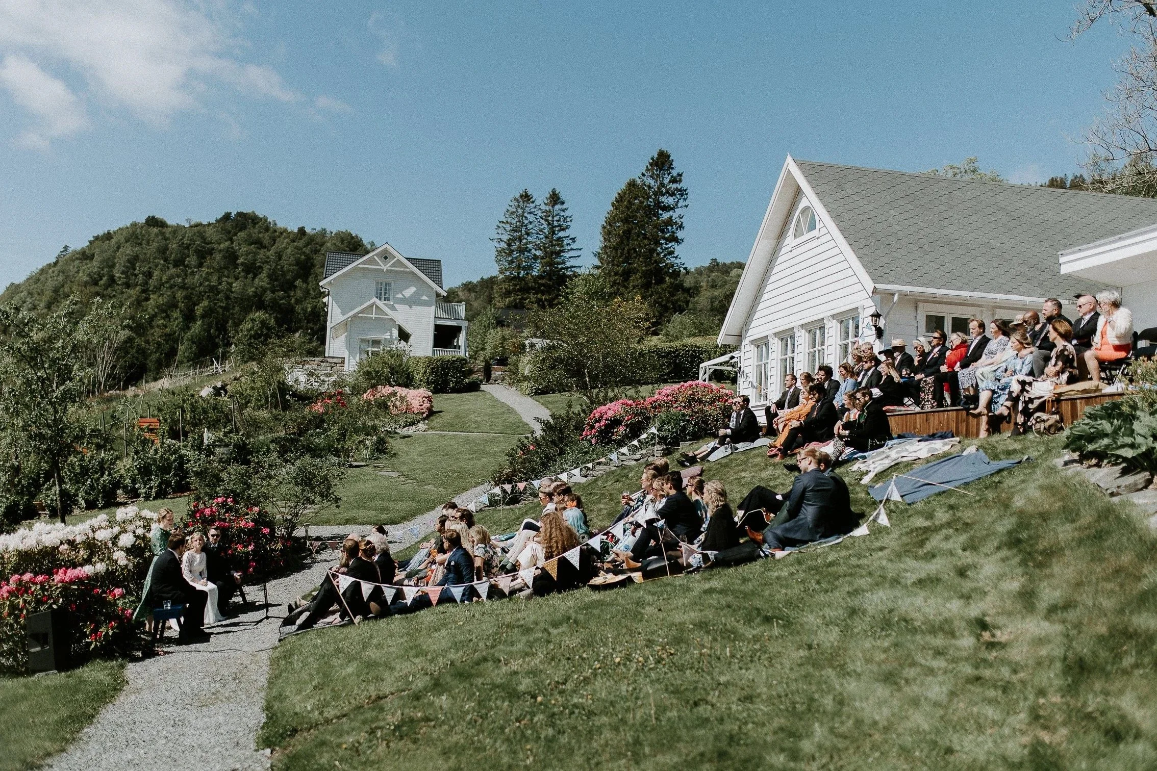 Utendørs bryllupsseremoni med gjester sittende i en gressbakke foran et hvitt hus, på en solrik dag med blå himmel, omgitt av blomster og trær.